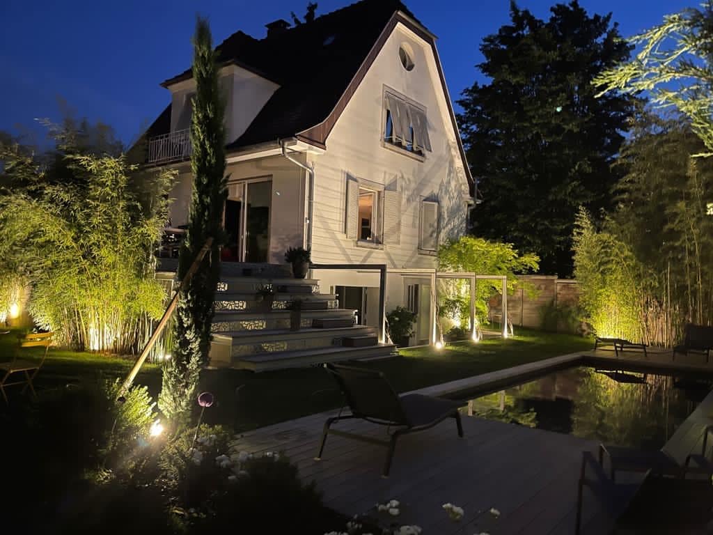 Élégante maison blanche illuminée la nuit. Jardin luxuriant avec bambous, escalier, piscine et terrasse en bois aménagée.