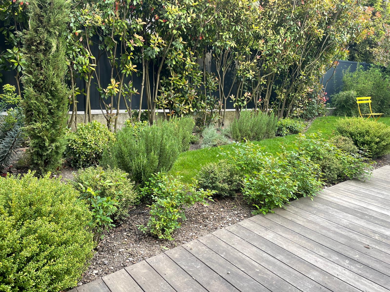 Jardin verdoyant avec terrasse en bois, buissons denses, romarin et haie d'arbustes feuillus. Une chaise jaune ressort au fond.