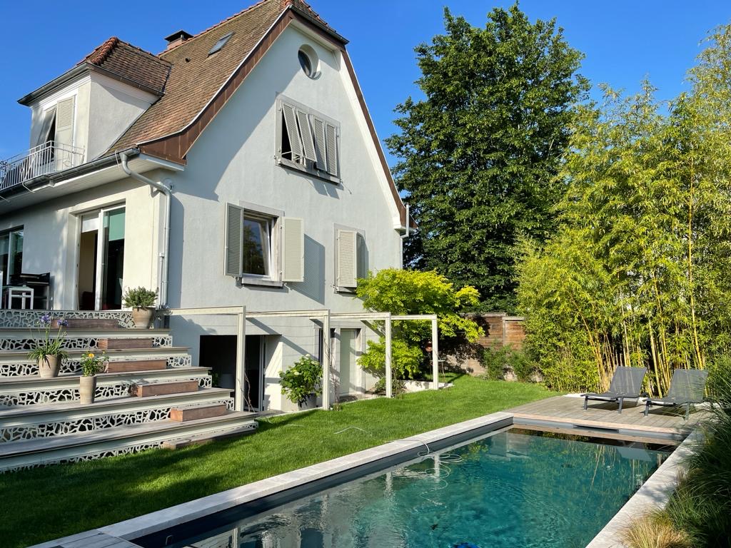 Élégante maison avec toit en tuiles, piscine moderne, pelouse verte et bambous. Des escaliers décoratifs mènent à une terrasse ensoleillée.