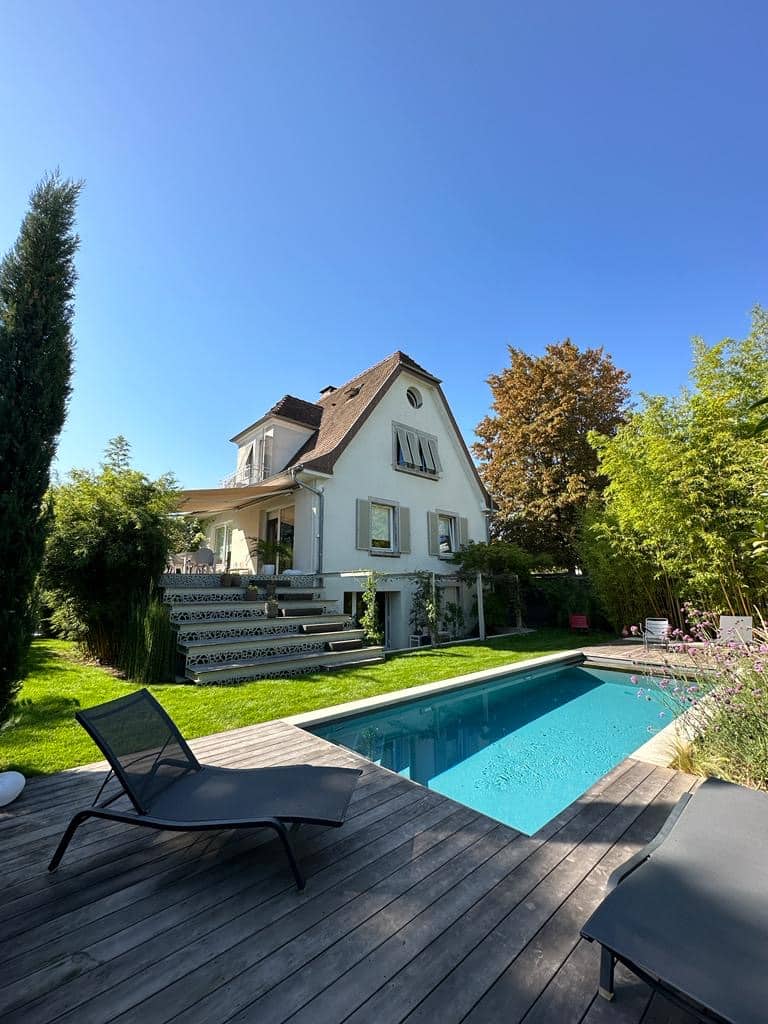 Grande maison beige avec toit en tuiles, piscine bleue et terrasse en bois. Jardin luxuriant, chaises longues sous un ciel bleu ensoleillé.