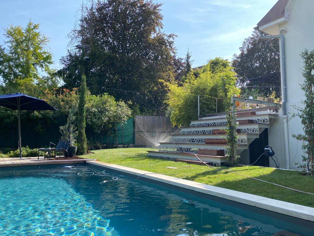 Piscine bleue, pelouse verte arrosée et escalier design menant à une terrasse ensoleillée. Maison moderne avec jardin luxuriant en été.
