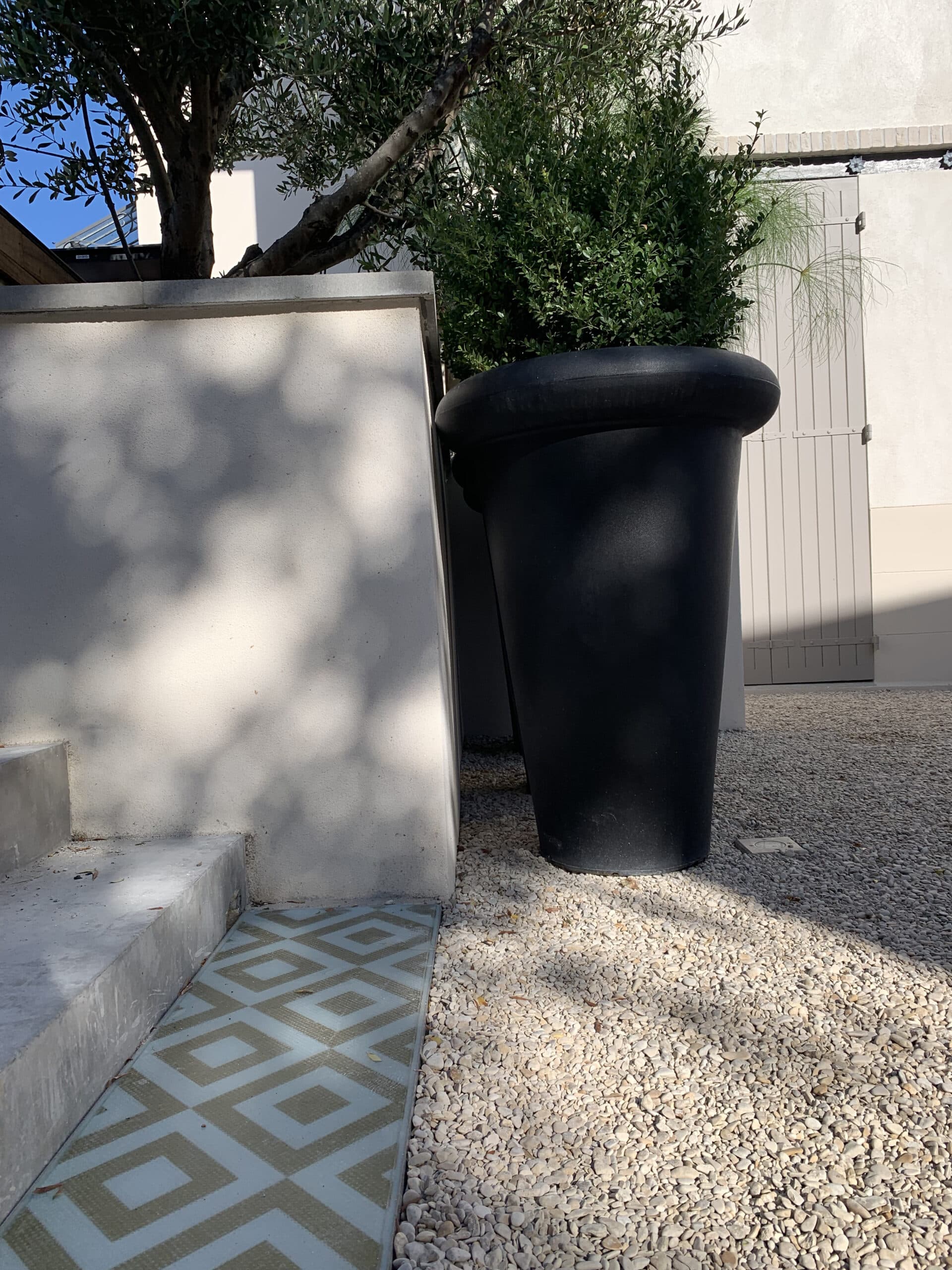Aménagement extérieur ensoleillé: gravier, escalier béton, carrelage géométrique bleu, grand pot noir, olivier, mur blanc ombragé.