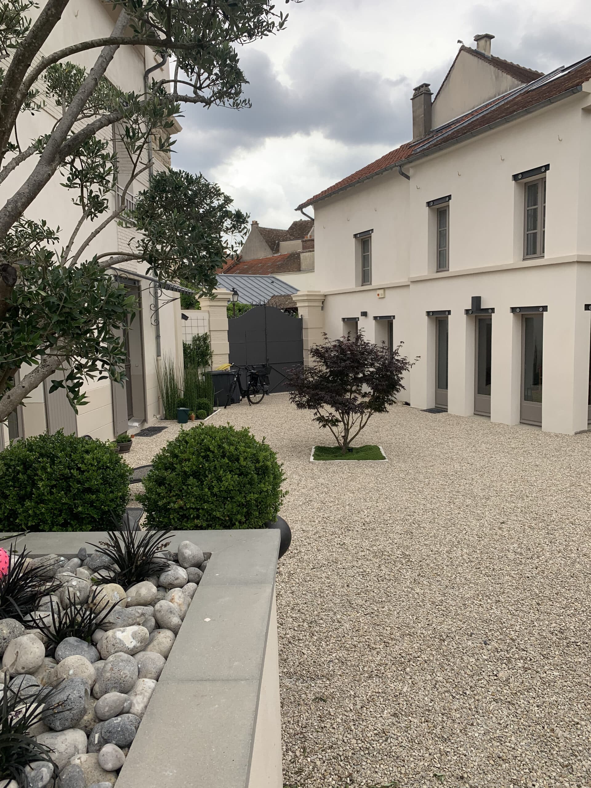 Cour moderne avec gravier beige clair, bâtiments blancs épurés, portail noir et verdure. Parterre de galets, plantes foncées et buissons verts en premier plan.