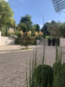 Allée de gravier clair, jardin en terrasses avec cyprès élancés, graminées dorées, arbustes et prêles ensoleillées. Ciel bleu.