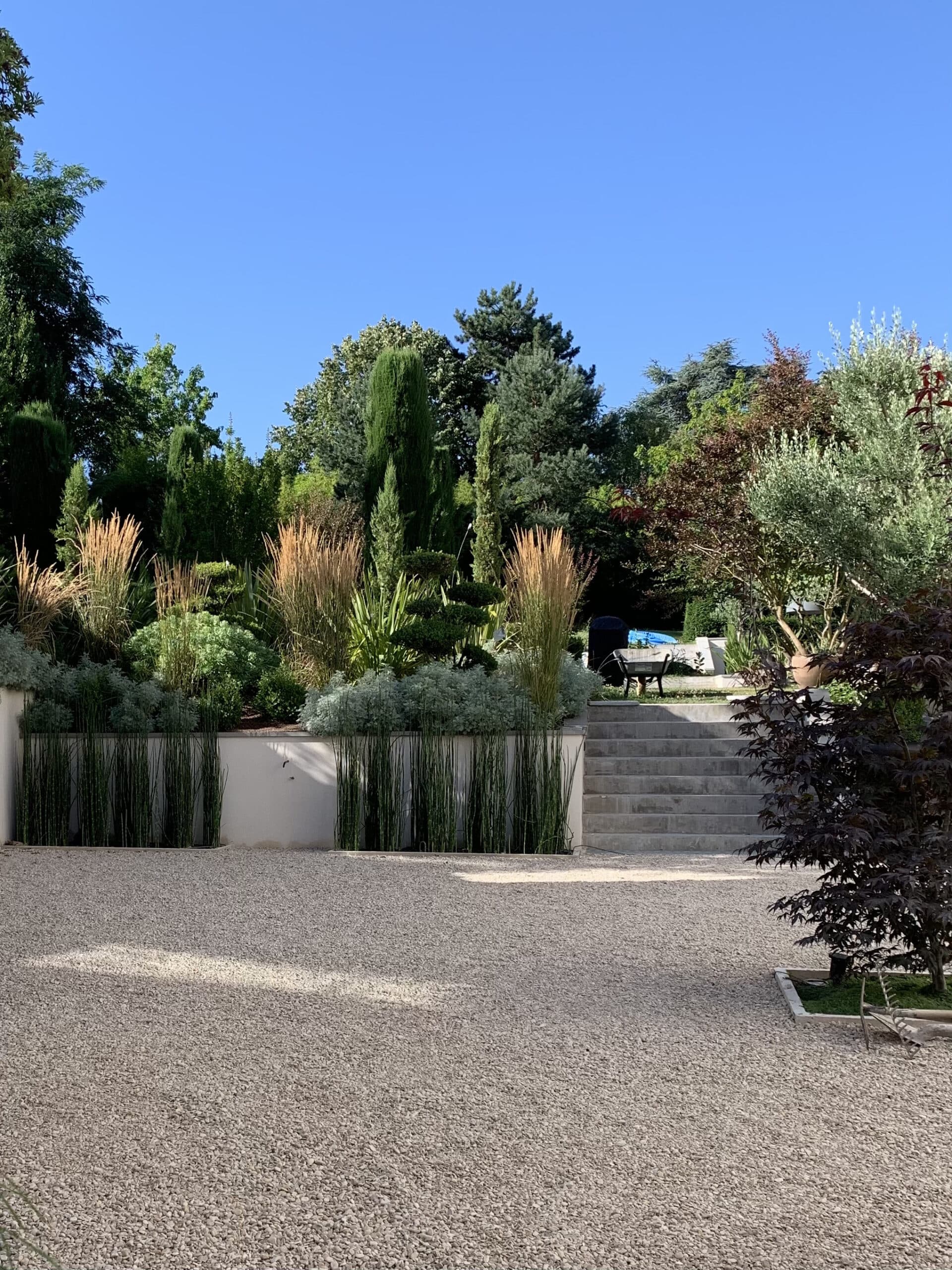 Grand jardin paysager avec chemin de gravier, mur blanc, graminées, arbres et escalier en béton sous un ciel bleu clair.