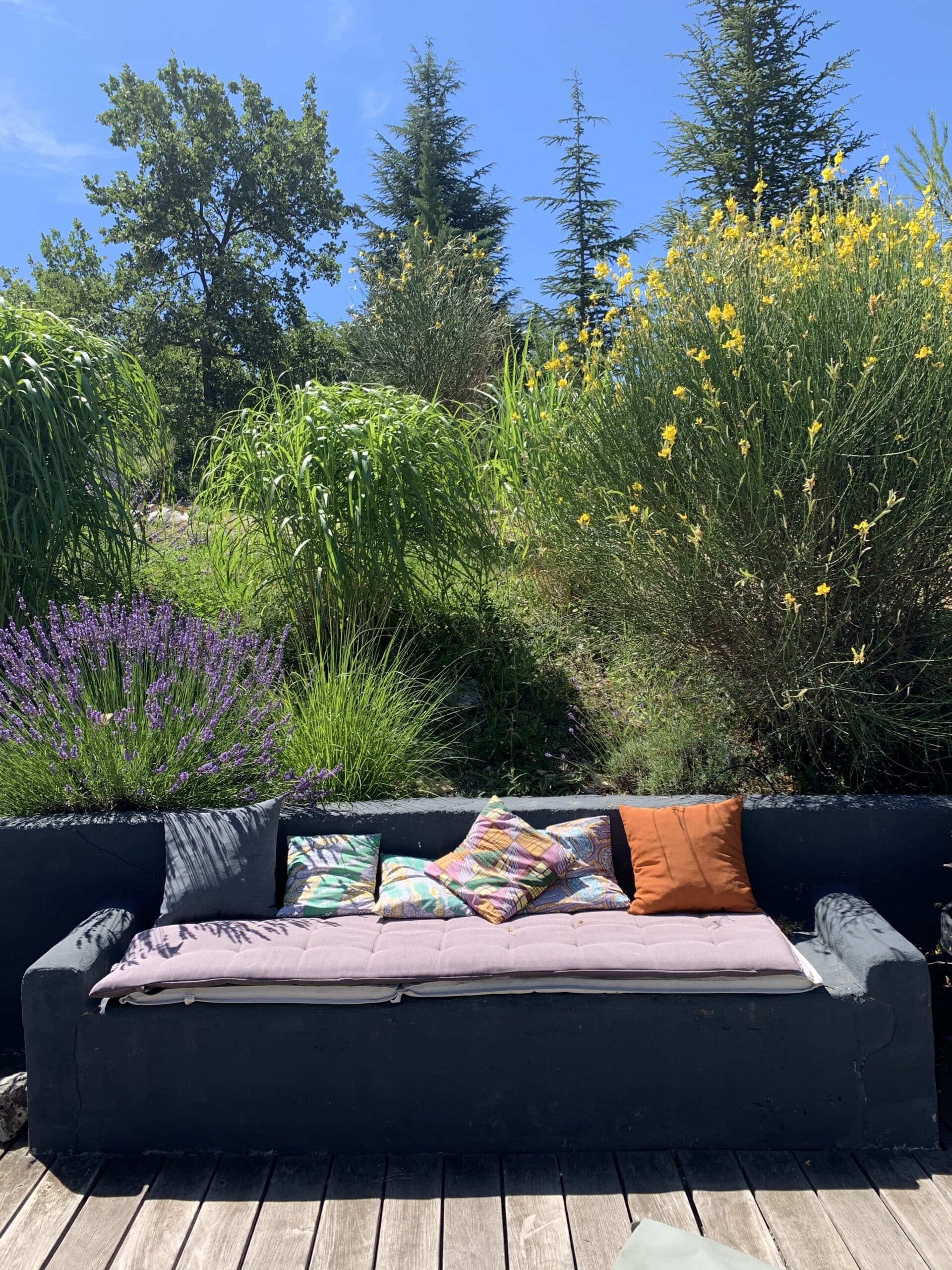 Banc extérieur gris avec coussins colorés sur terrasse en bois. Jardin méditerranéen luxuriant: lavande violette, genêt jaune, arbres, ciel bleu.