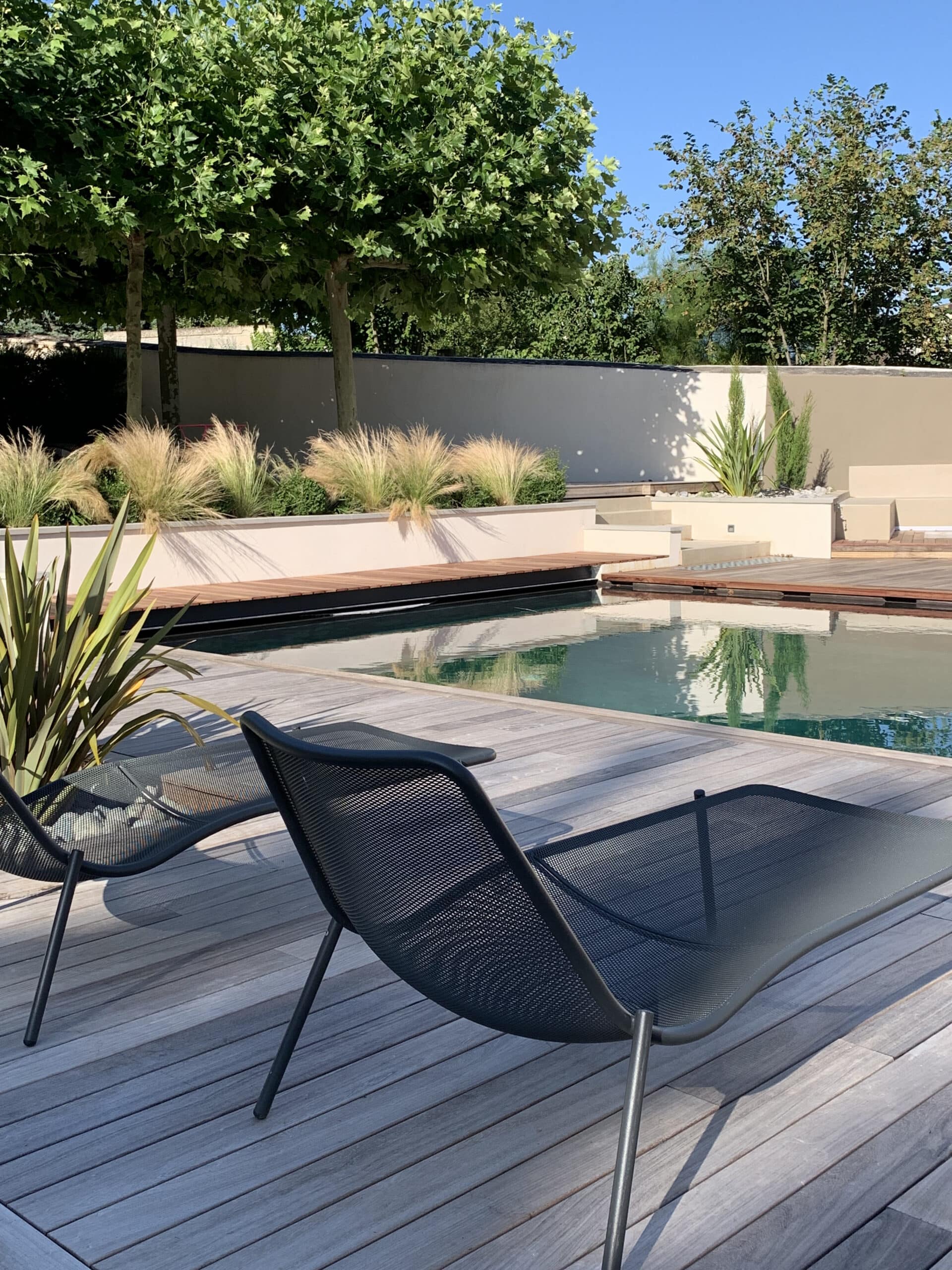Piscine moderne et terrasse en bois avec deux chaises longues. Jardin paysager luxuriant, arbres et graminées ornementales, murs clairs, ciel bleu.