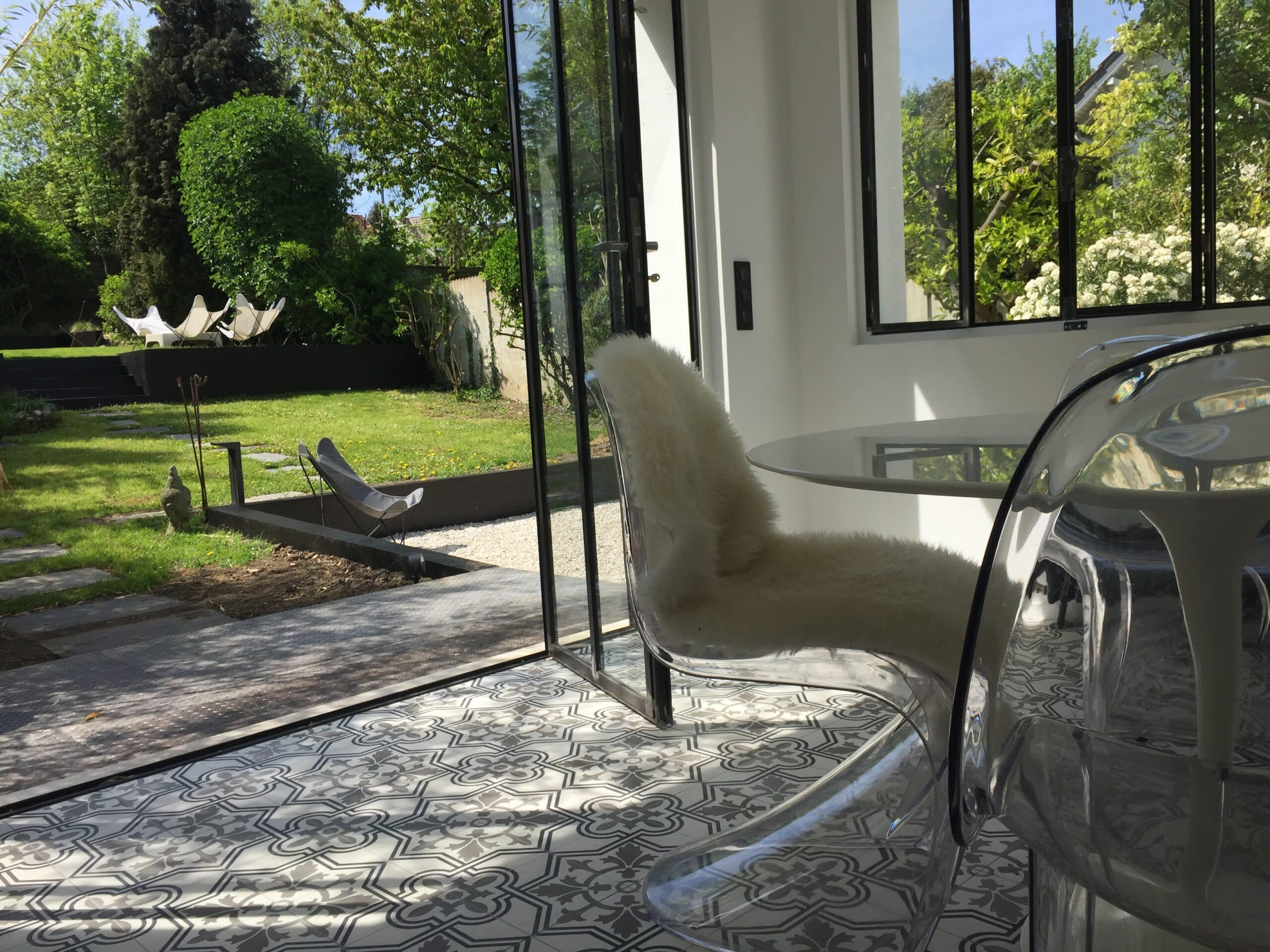 Salon moderne lumineux avec carrelage motifs, table en verre, chaises transparentes. Vue sur jardin luxuriant, transats et statue de Bouddha.