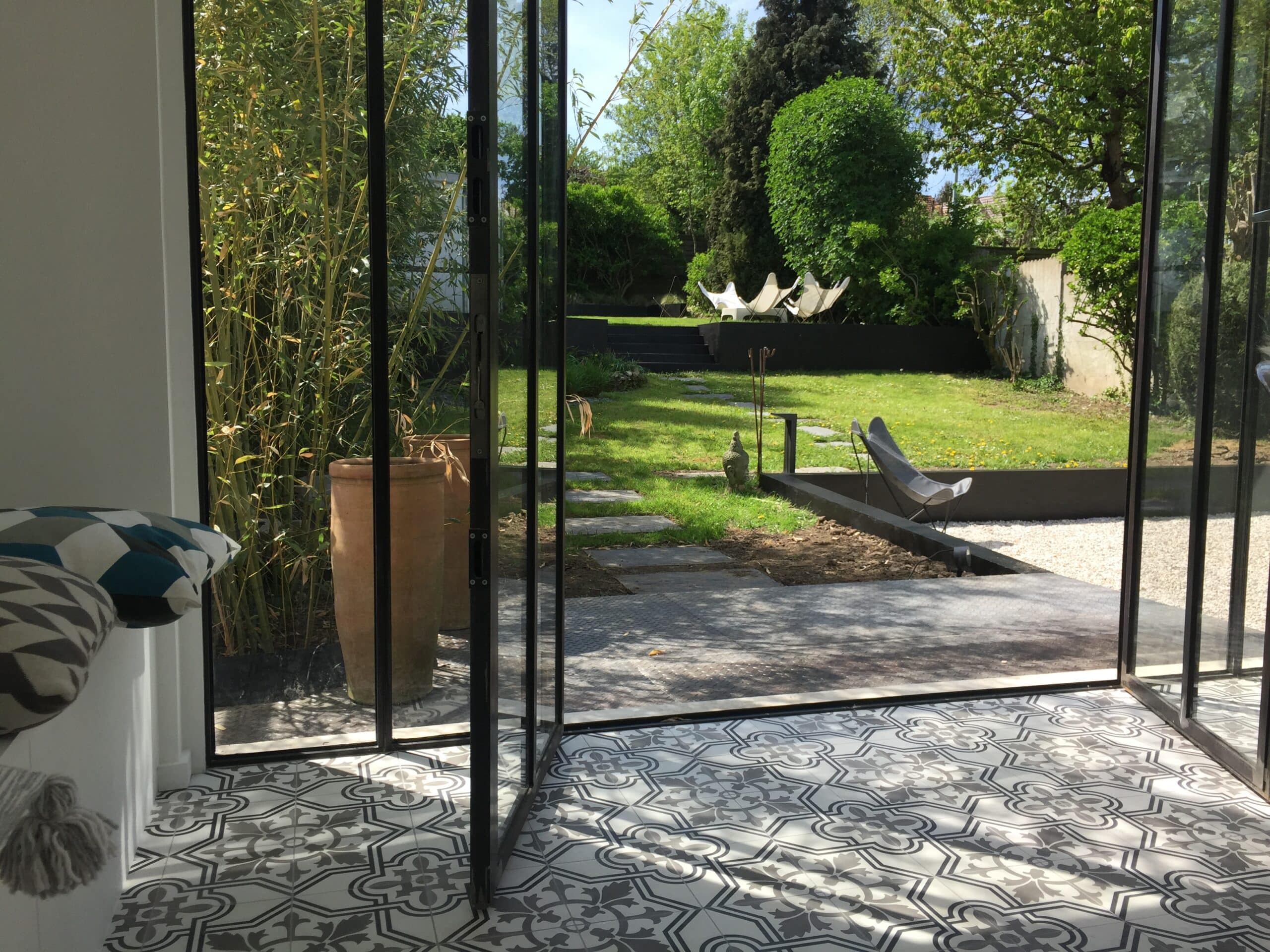 Luminosité d'un jardin luxuriant avec bambous, pelouse et terrasse, visible à travers des portes vitrées depuis une pièce aux carreaux ciment motifs gris.