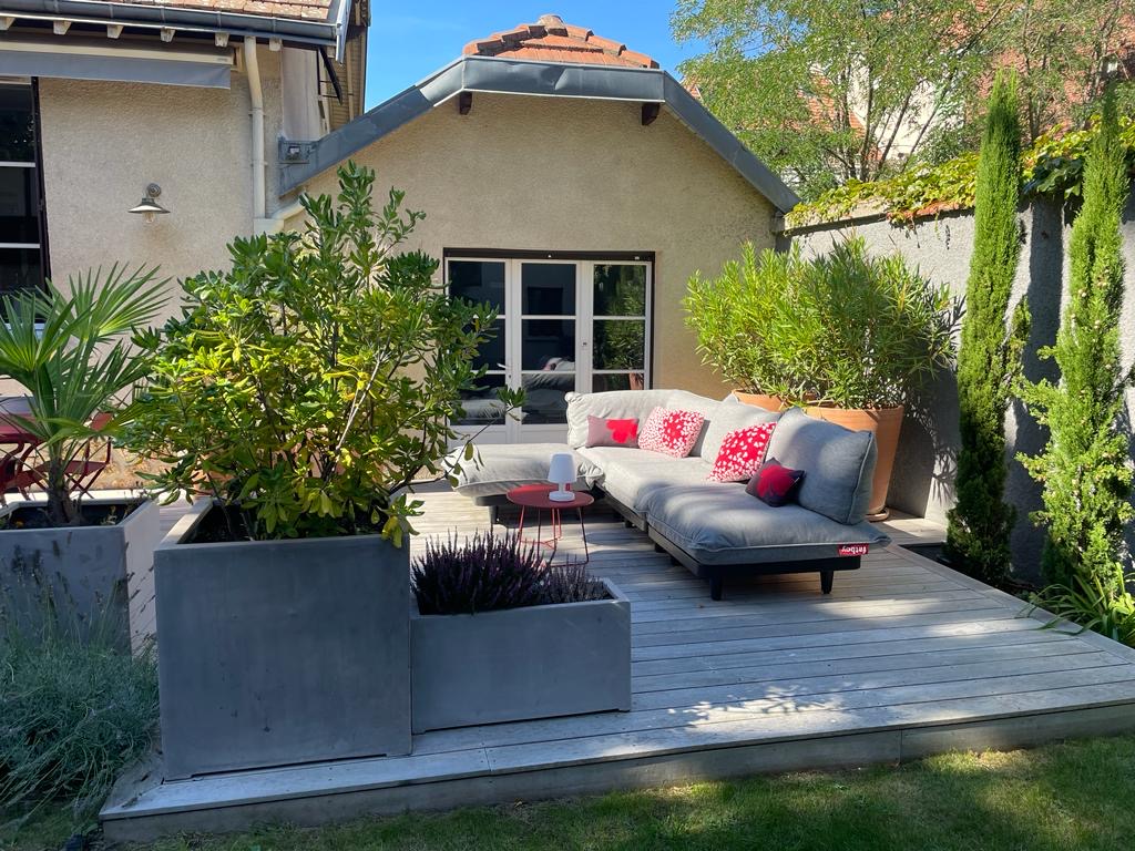 Terrasse en bois avec salon de jardin modulable gris Fatboy, coussins rouges. Plantes en pots, jardinières béton, mur de maison. Jour ensoleillé.