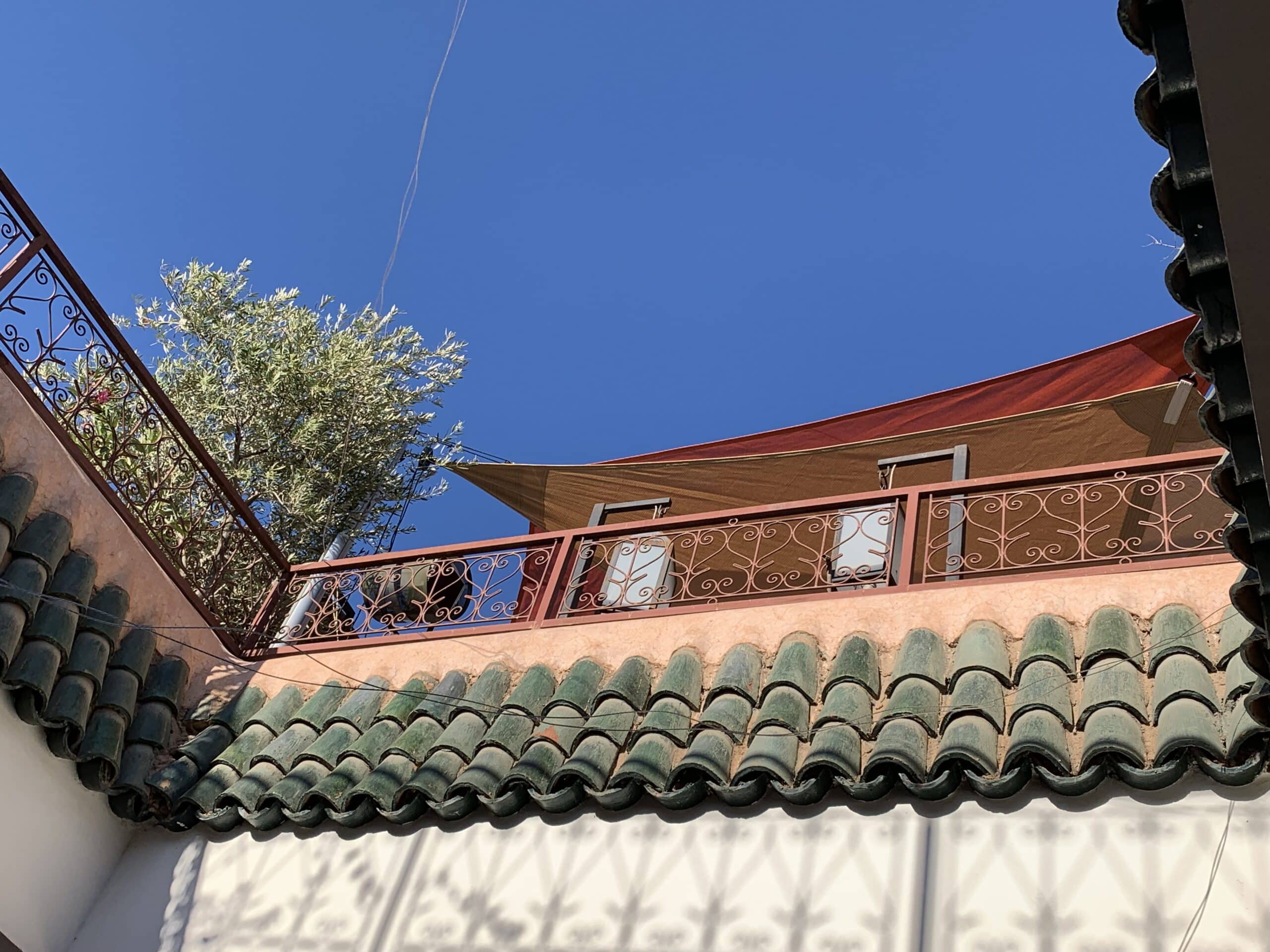Vue de bas en haut d'une façade marocaine avec toits verts, balcon en fer forgé orné, ciel bleu vif, un olivier et un auvent rouge.