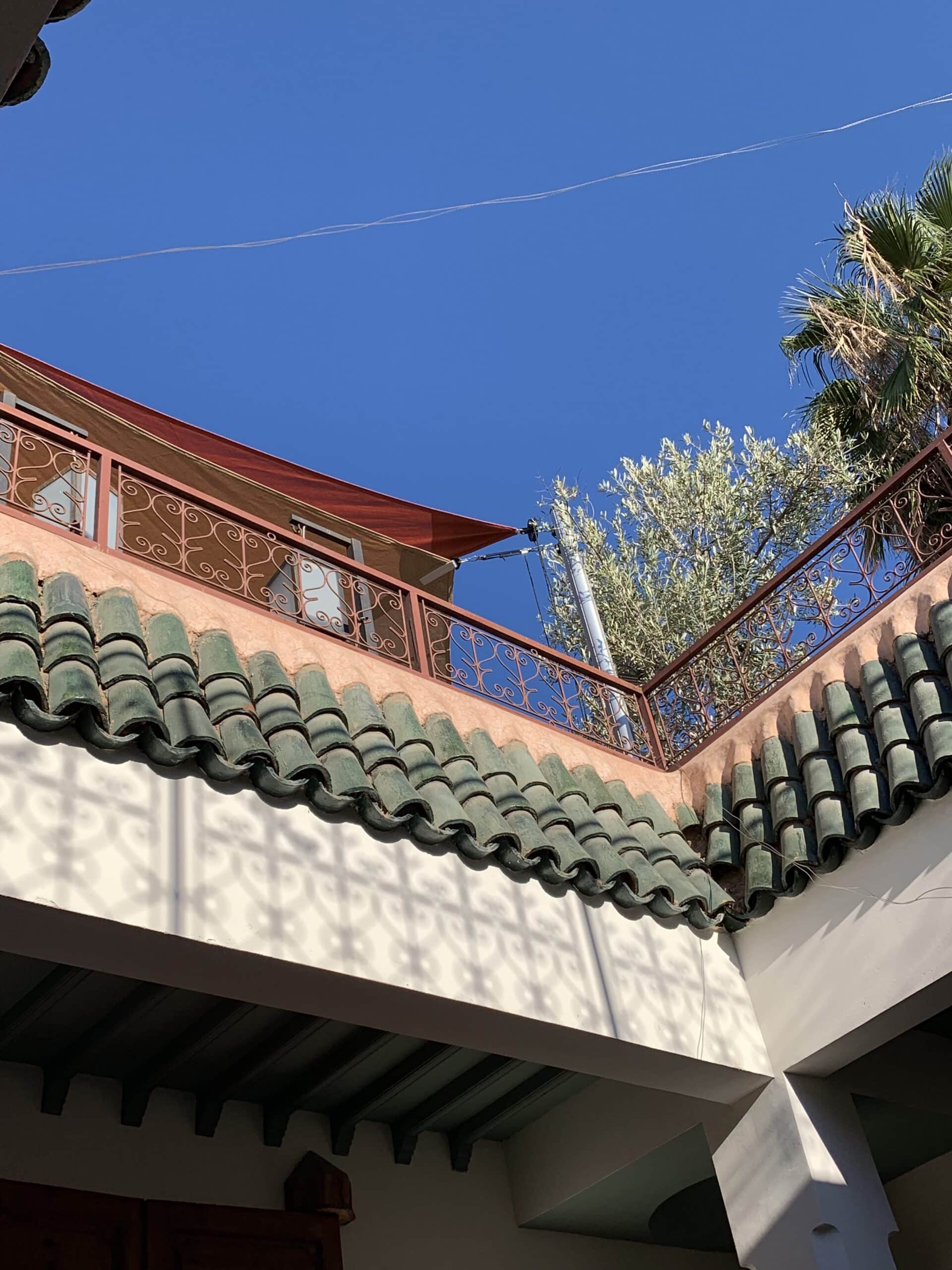 Architecture marocaine. Tuiles vertes, mur terre cuite, garde-corps fer forgé. Ciel bleu, palmier, ombres motifs sur mur blanc.