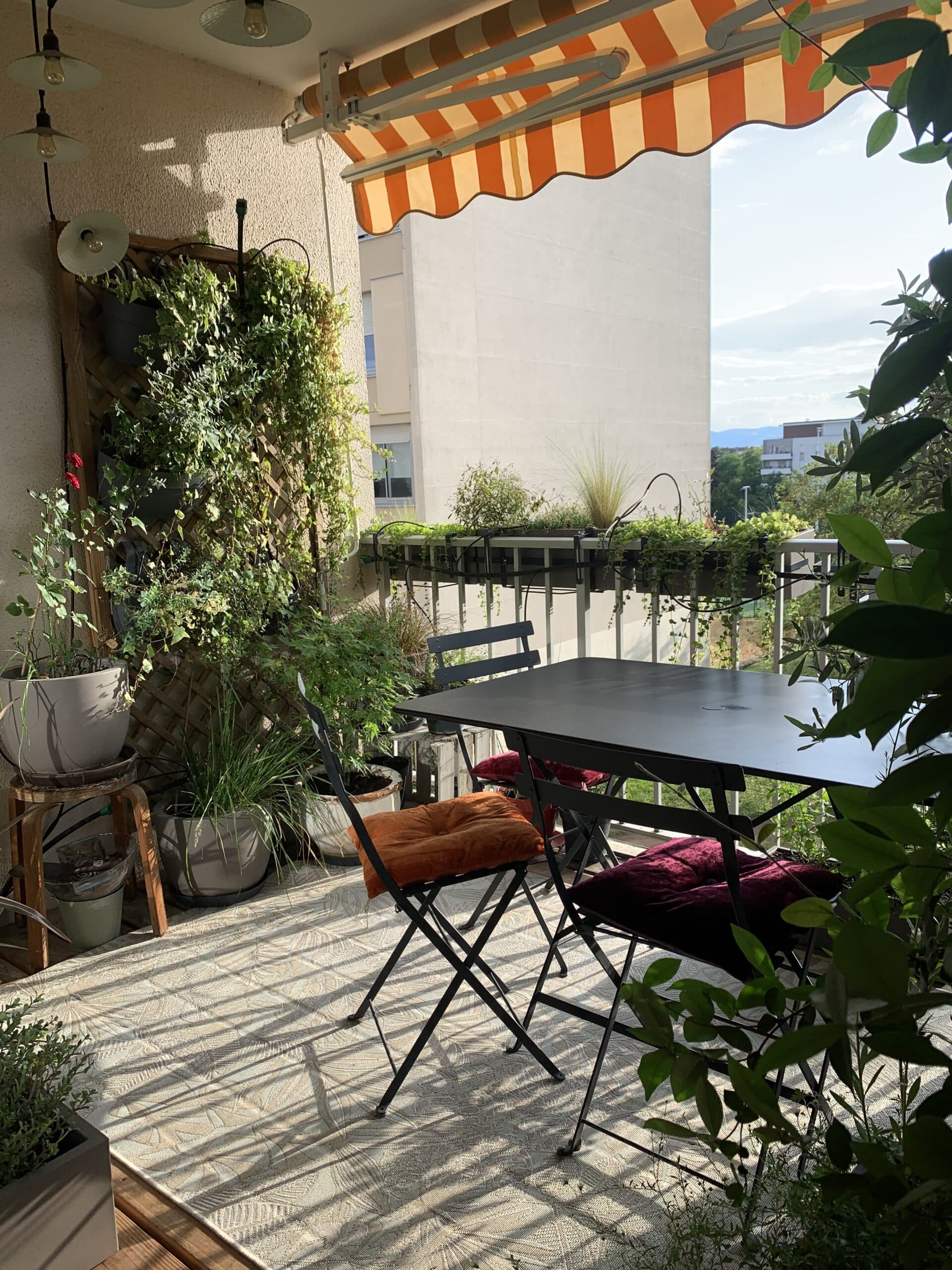 Balcon ensoleillé avec jardin vertical, table noire et chaises (coussins orange/grenat), store rayé orange. Vue urbaine.