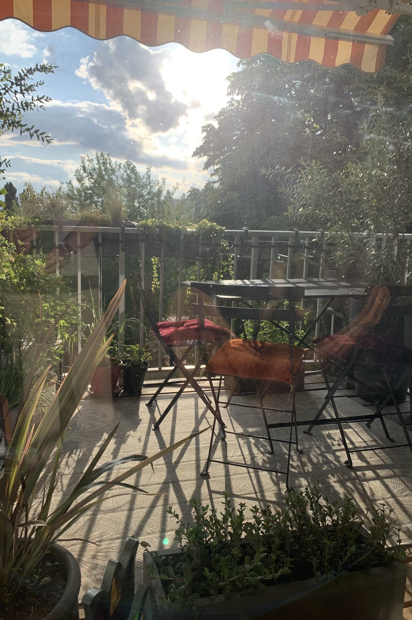 Balcon baigné de soleil avec auvent rayé, table, chaises aux coussins colorés, et plantes. Longues ombres sur le sol, lumière éclatante.