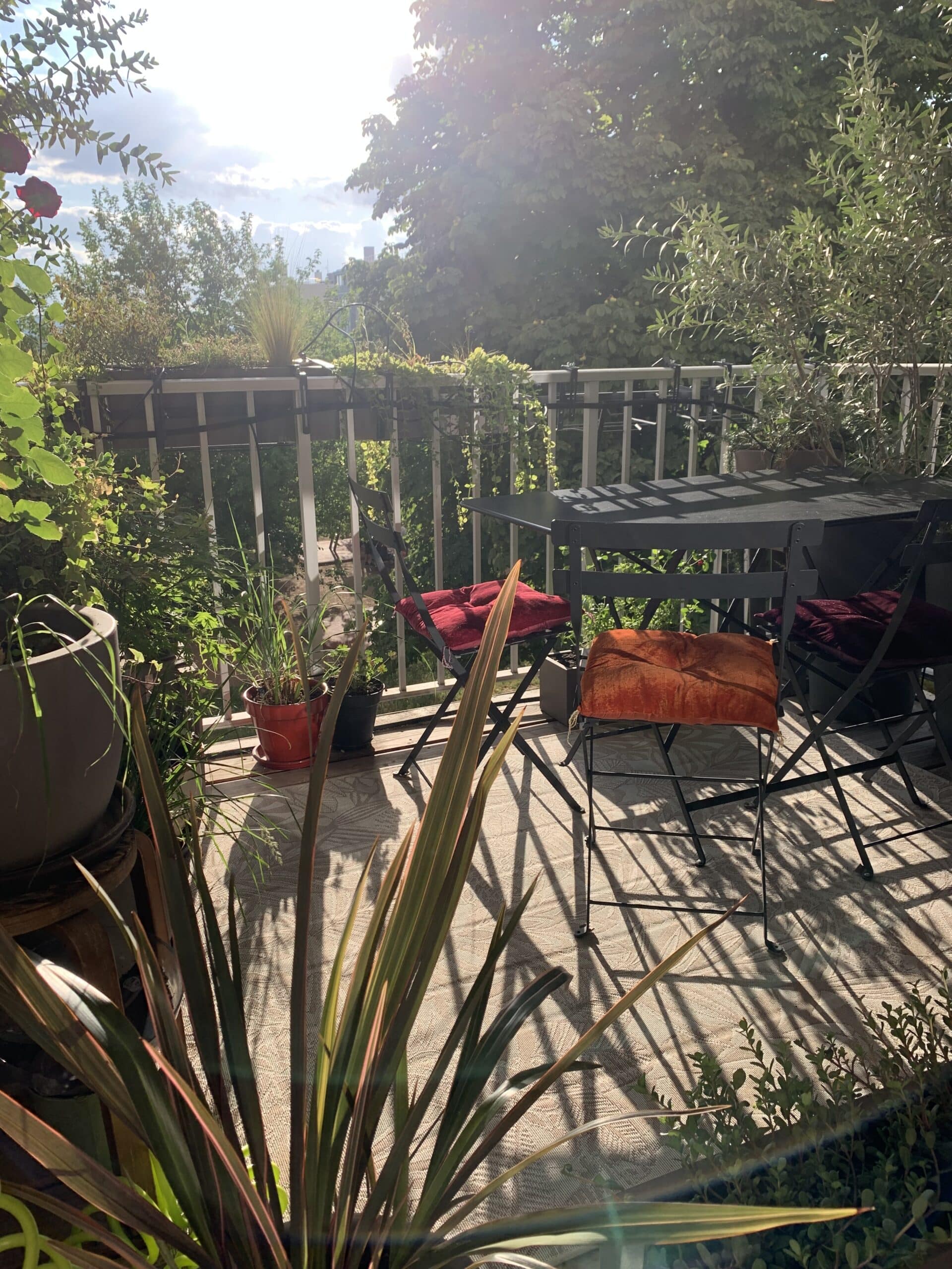 Balcon ensoleillé avec table et chaises cosy aux coussins rouges, entouré de plantes luxuriantes. Les ombres du soleil s'allongent sur le sol.