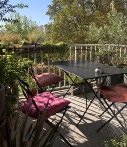 Balcon fleuri avec table noire pliante et trois chaises rembourrées aux coussins colorés, éclairé par un fort soleil créant des ombres graphiques.