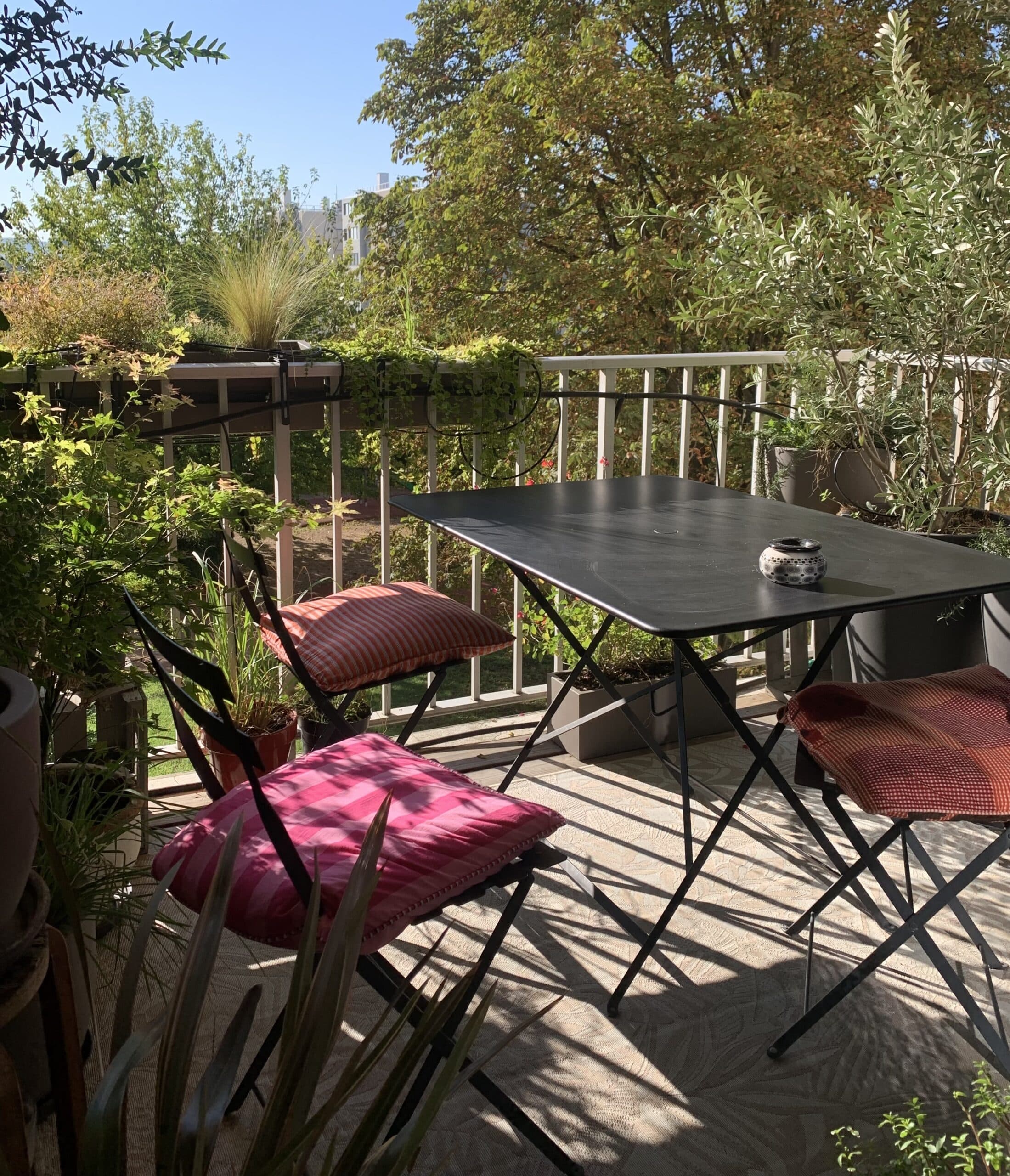 Balcon fleuri avec table noire pliante et trois chaises rembourrées aux coussins colorés, éclairé par un fort soleil créant des ombres graphiques.