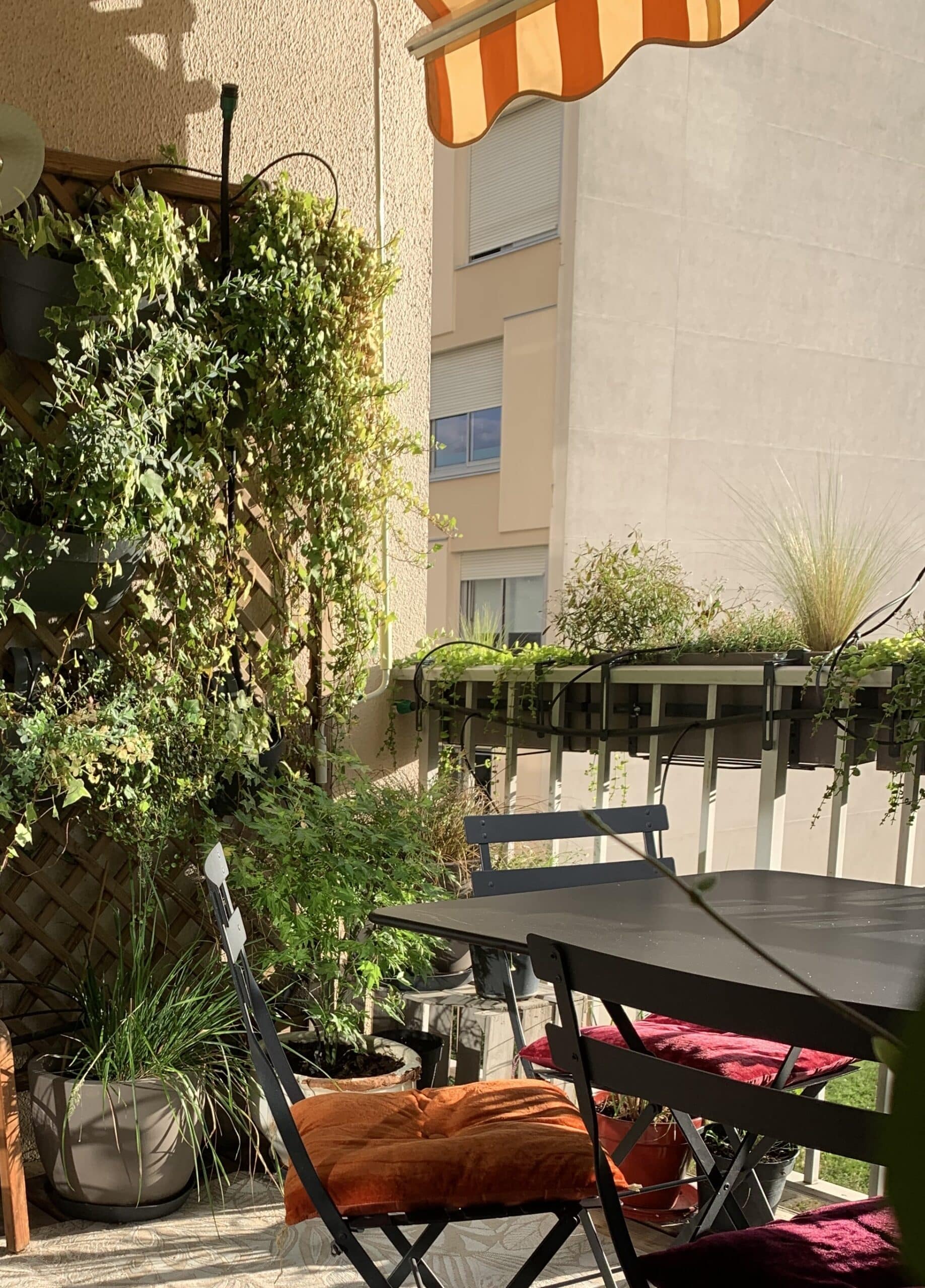 Balcon ensoleillé avec jardin vertical verdoyant, table et chaises aux coussins orange et rouges. Ambiance végétale urbaine et cosy.