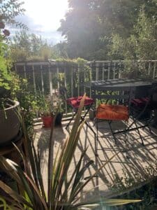 Balcon ensoleillé avec mobilier de jardin noir, coussins rouge et orange, et abondance de plantes créant des ombres graphiques.