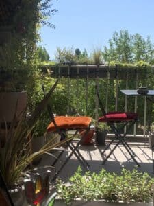 Balcon ensoleillé avec deux chaises pliantes, une orange et une rouge, entourées de plantes vertes luxuriantes sous un ciel bleu clair.