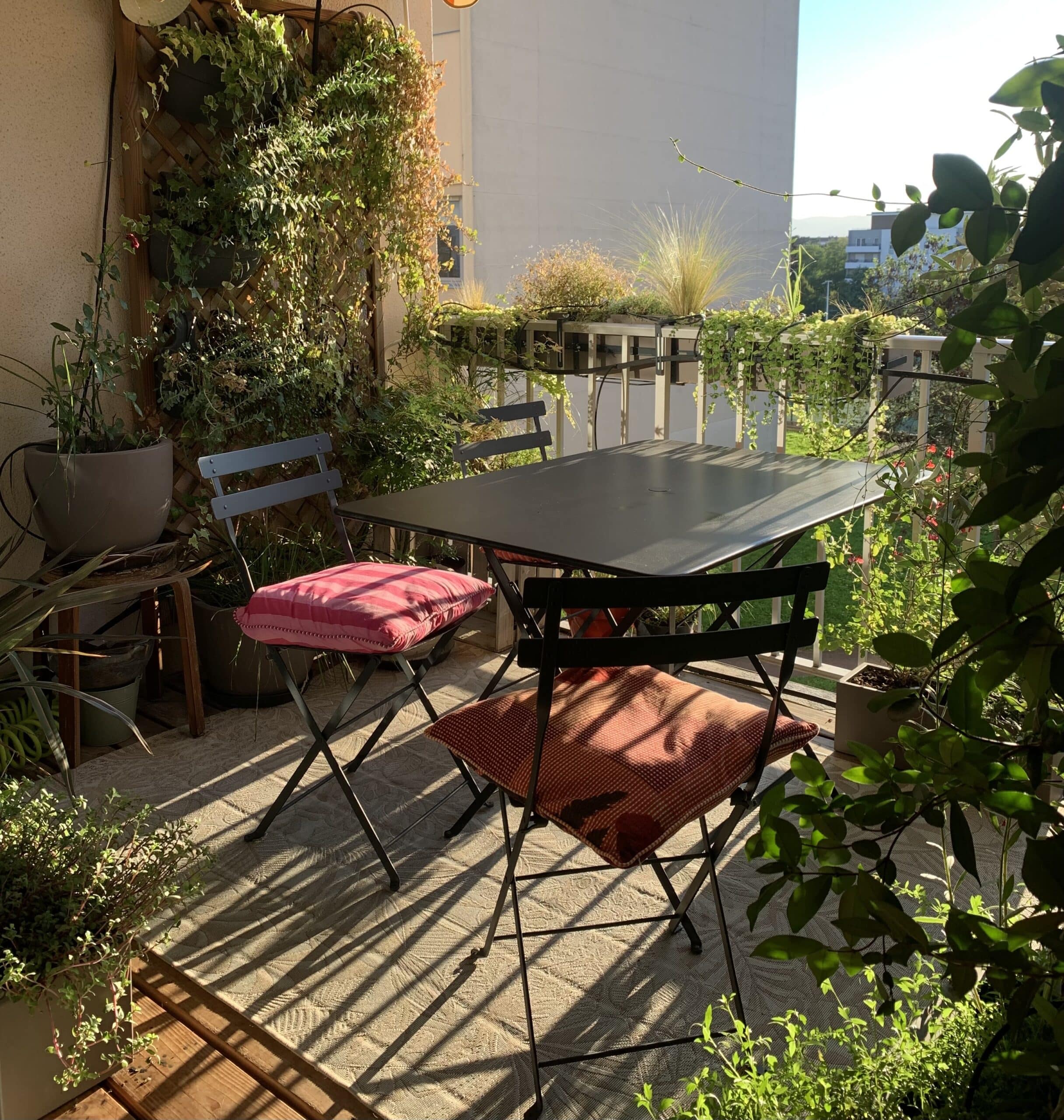 Balcon fleuri baigné de soleil avec table noire et deux chaises pliantes ornées de coussins rouge/rose, ombres rayées sur le sol.