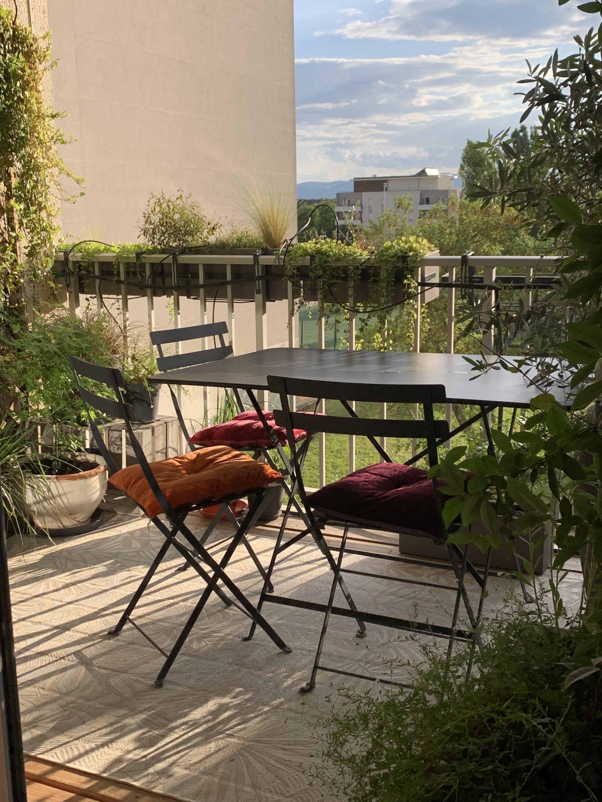 Balcon ensoleillé avec table, chaises, coussins colorés et plantes vertes luxuriantes. Ombres nettes sur le carrelage. Vue sur la ville et le ciel.