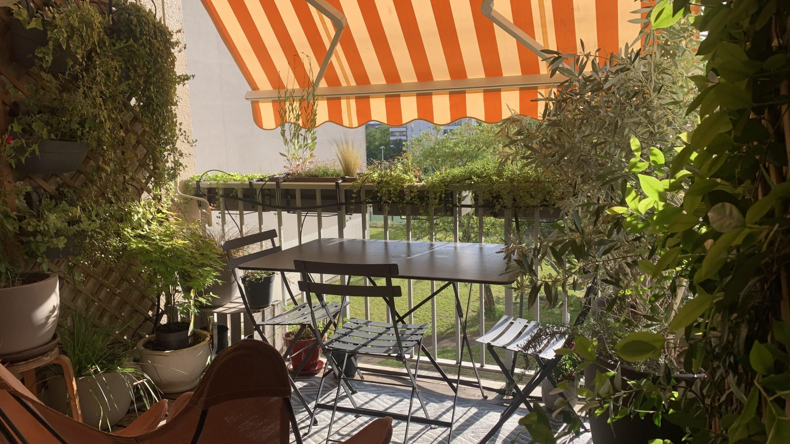 Balcon ensoleillé avec auvent rayé orange et blanc, table et chaises grises, et de nombreuses plantes vertes luxuriantes.