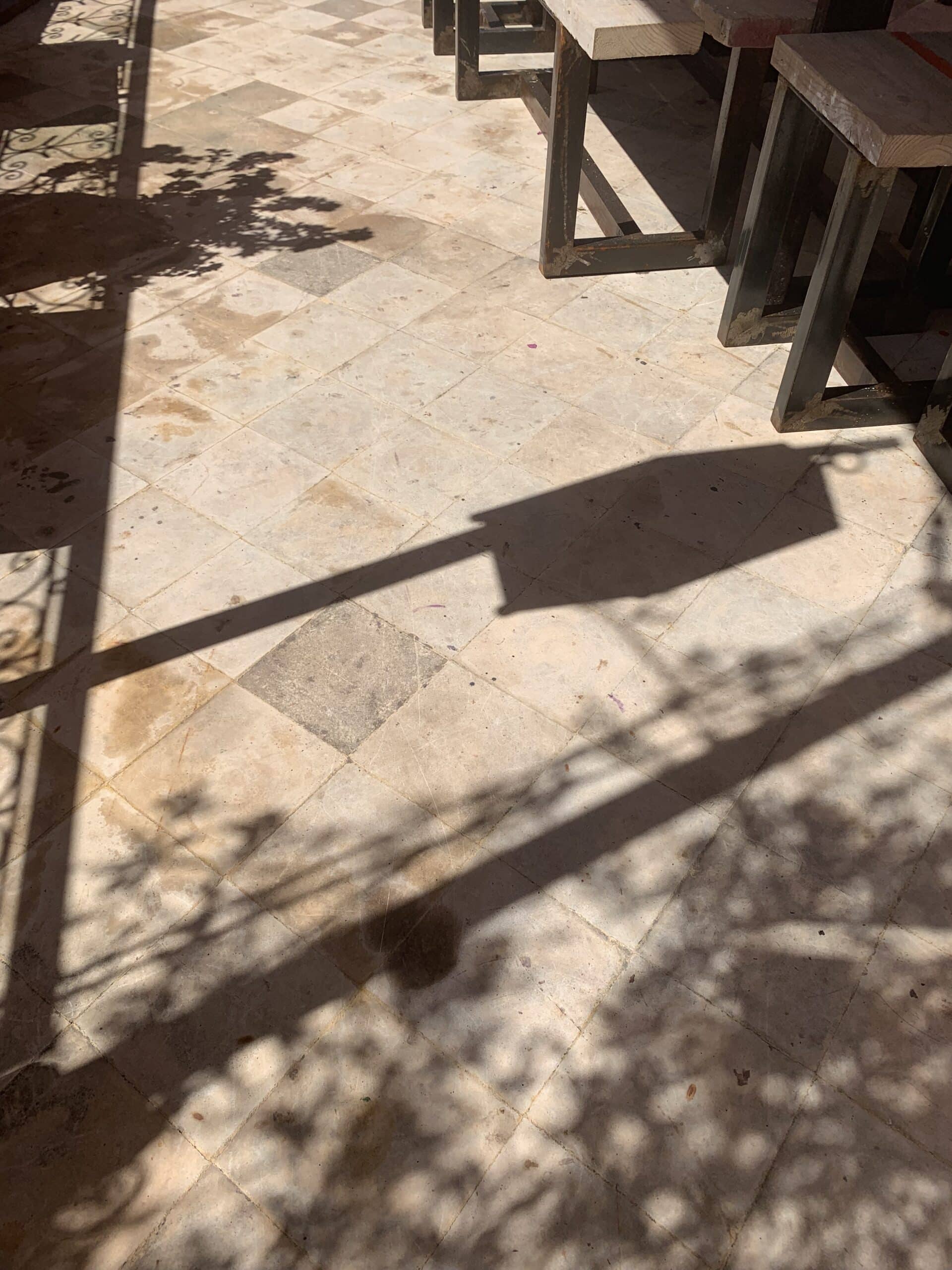 Carrelage clair d'un patio ensoleillé, ombres de tables en bois et métal, et motifs végétaux d'une balustrade.