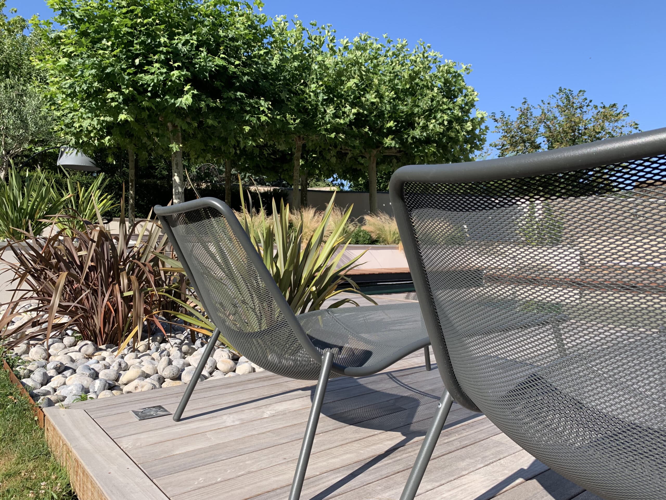 Terrasse bois moderne avec deux chaises de jardin gris, entourée de rochers, plantes luxuriantes et grands arbres verts. Ciel bleu éclatant.