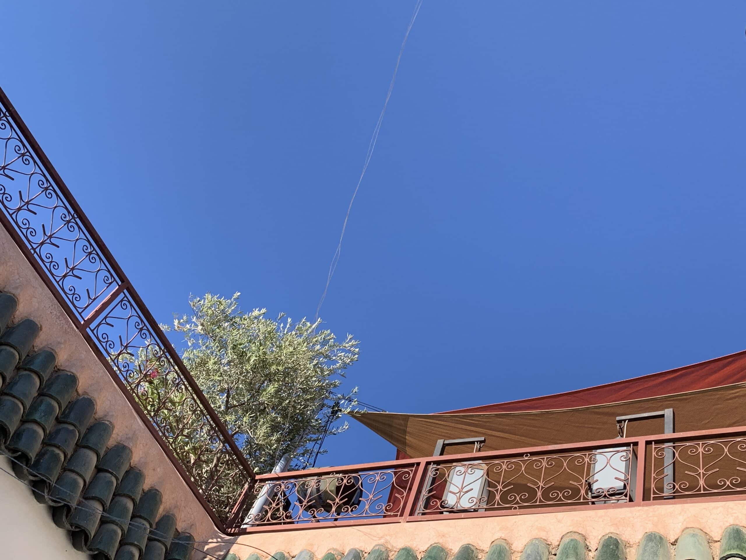 Ciel bleu azur sur toit terrasse ensoleillé. Garde-corps en fer forgé, tuiles vertes et voile d'ombrage. Un arbre se dresse derrière.