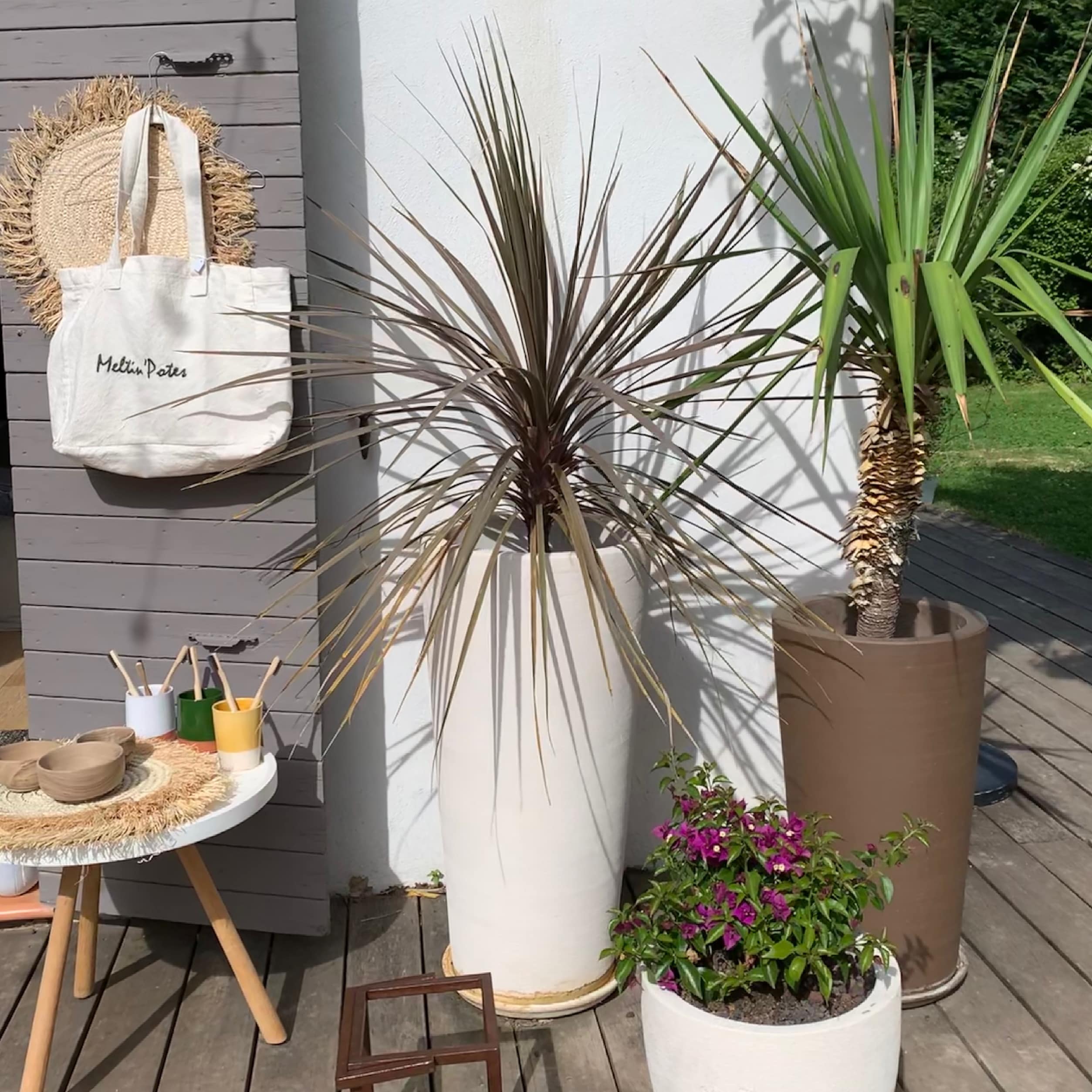 Sac 'Meltin'Potes', chapeau paille, bols bois et brosses bambou sur table. Grandes plantes Dracaena, Yucca, Bougainvillier sur terrasse bois.