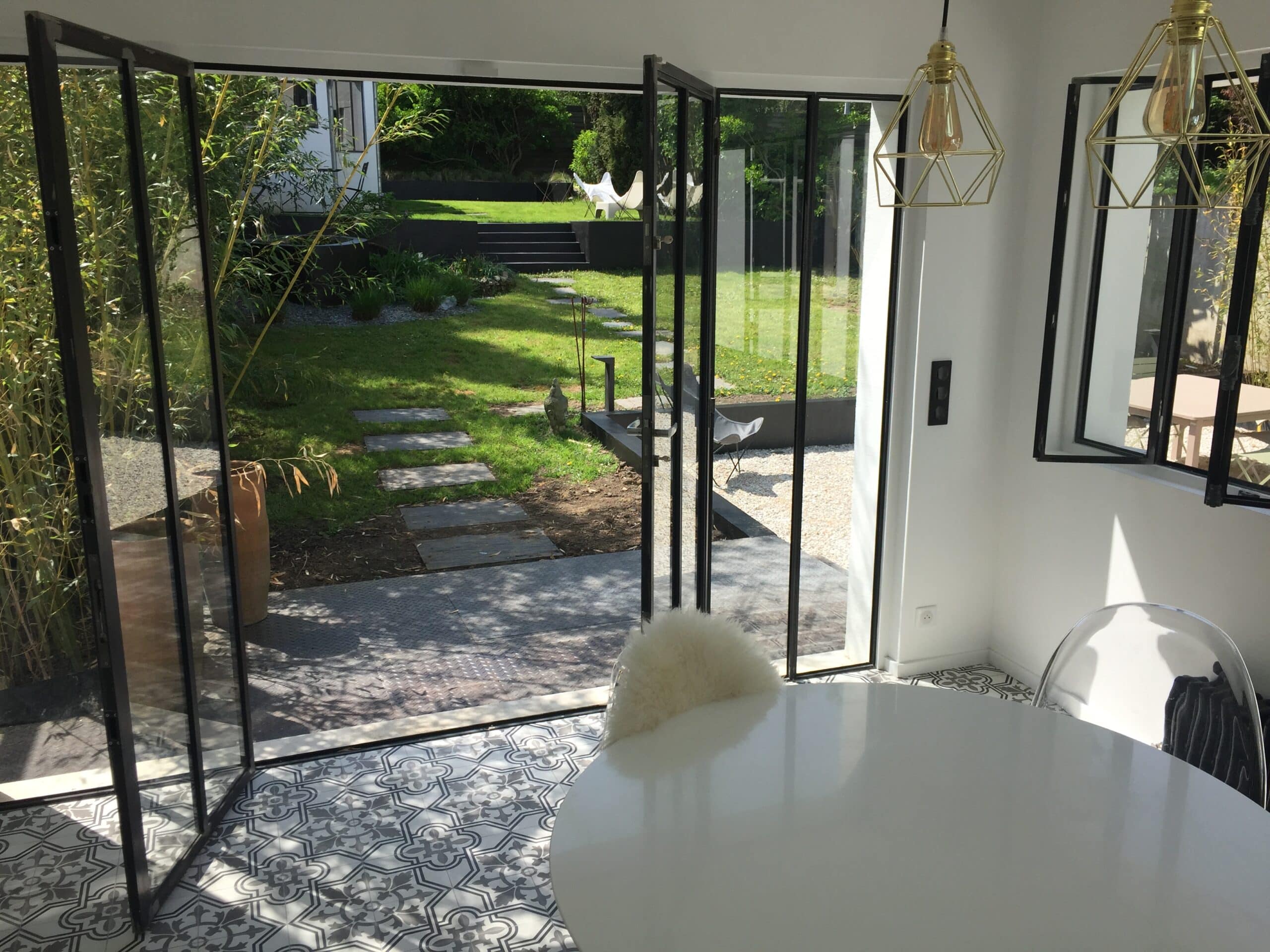 Intérieur moderne ouvrant sur jardin. Portes vitrées noires, carrelage graphique, table blanche et luminaires dorés. Jardin luxuriant avec pelouse, bambous et escaliers.