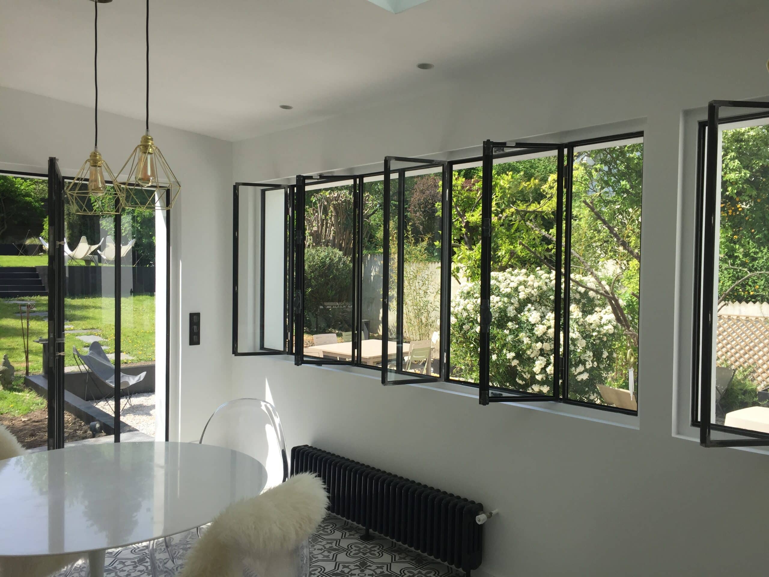 Salle à manger moderne et lumineuse. Table blanche, chaises design, radiateur noir. Baies vitrées ouvertes sur jardin verdoyant et ensoleillé.