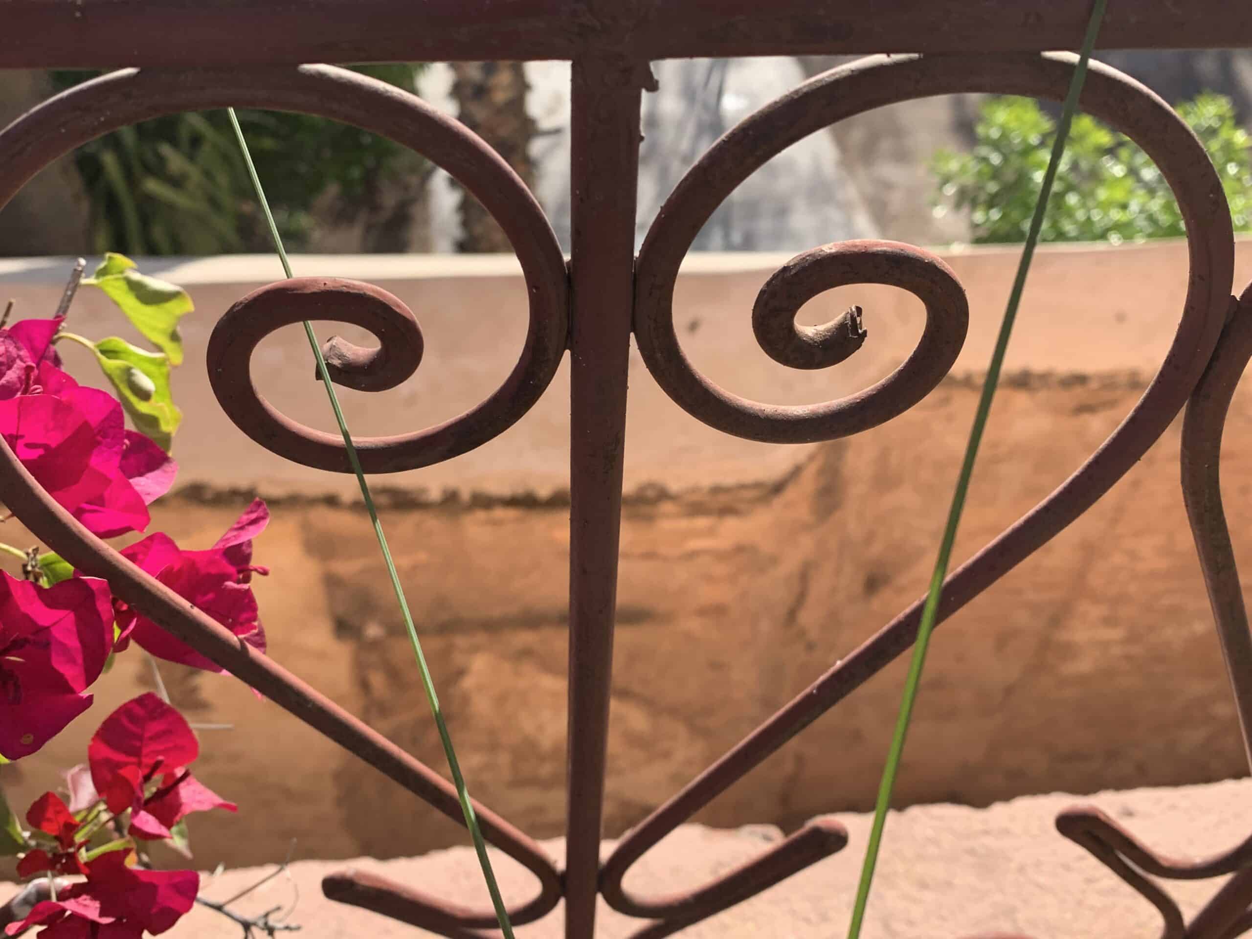 Ferronnerie rouille à motifs de cœurs et volutes, parsemée de bougainvilliers roses éclatants, devant un mur ocre flou.