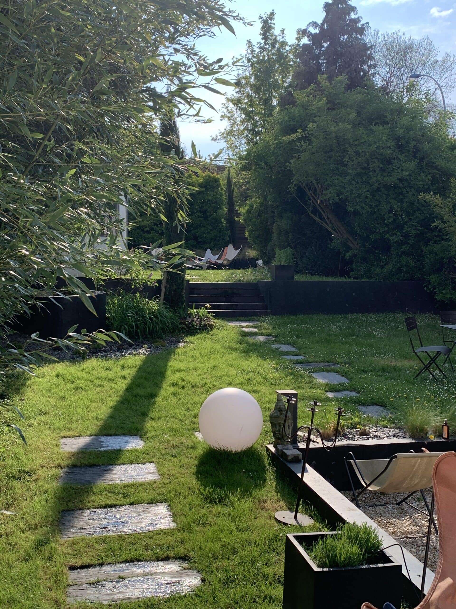 Jardin ensoleillé: pelouse verte, chemin d'ardoises brillantes, lampadaire sphérique, statue bouddha, chaises longues, arbres et bambous.