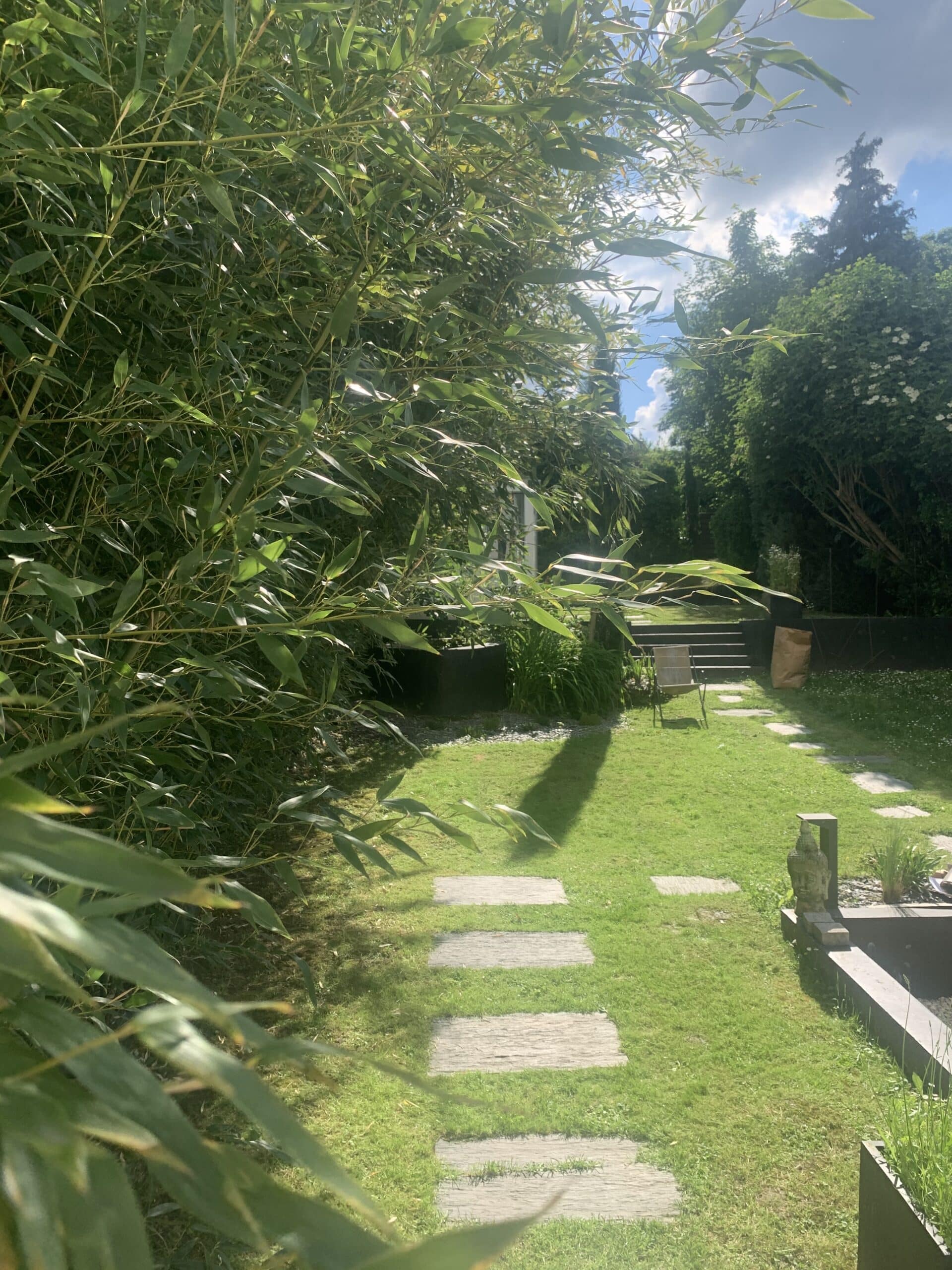 Jardin verdoyant et ensoleillé avec des bambous luxuriants, un chemin de dalles en pierre traversant la pelouse, et une statue.