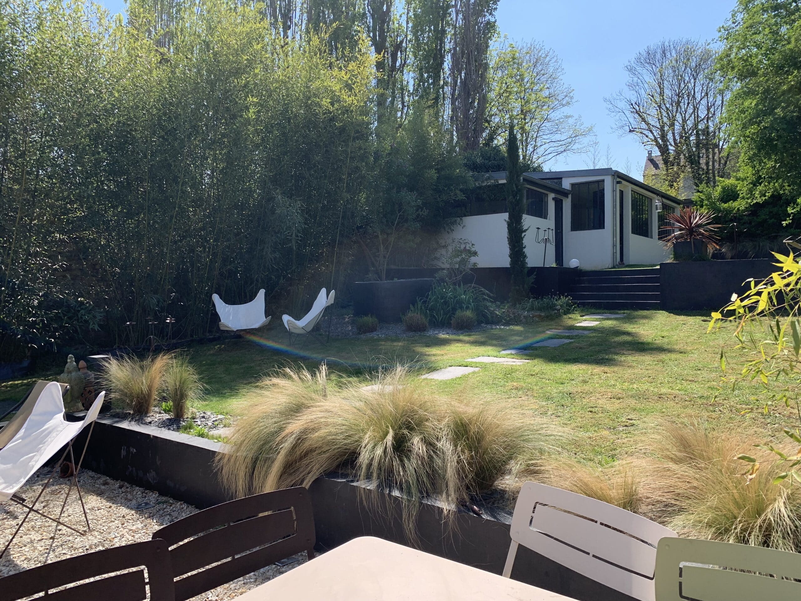 Jardin contemporain ensoleillé avec pelouse, terrasse, chaises, bambous luxuriants et maison moderne. Un petit arc-en-ciel apparaît sur l'herbe.