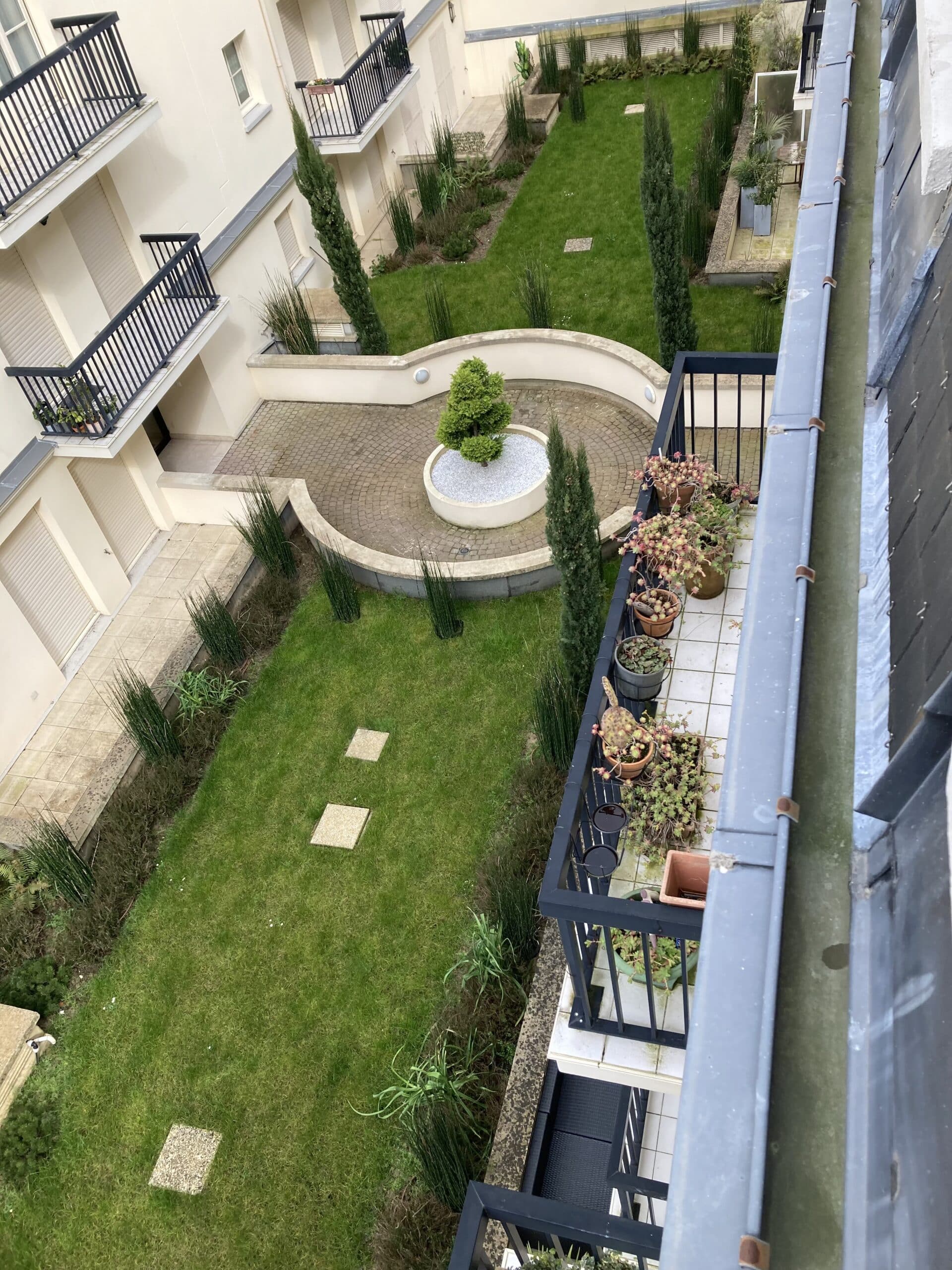 Jardin de cour d'immeuble vu d'en haut : pelouse, arbustes élancés, arbre central, allées pavées et balcons avec pots de fleurs.