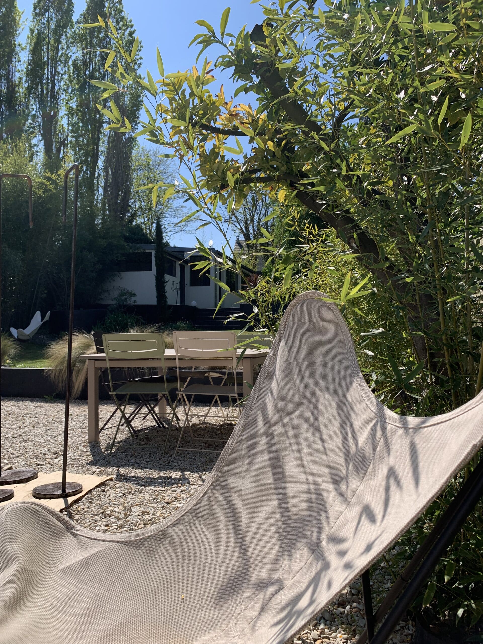 Chaise longue beige à ombres végétales, salon de jardin sur gravier. Verdure luxuriante, bambous et maison moderne sous ciel bleu ensoleillé.