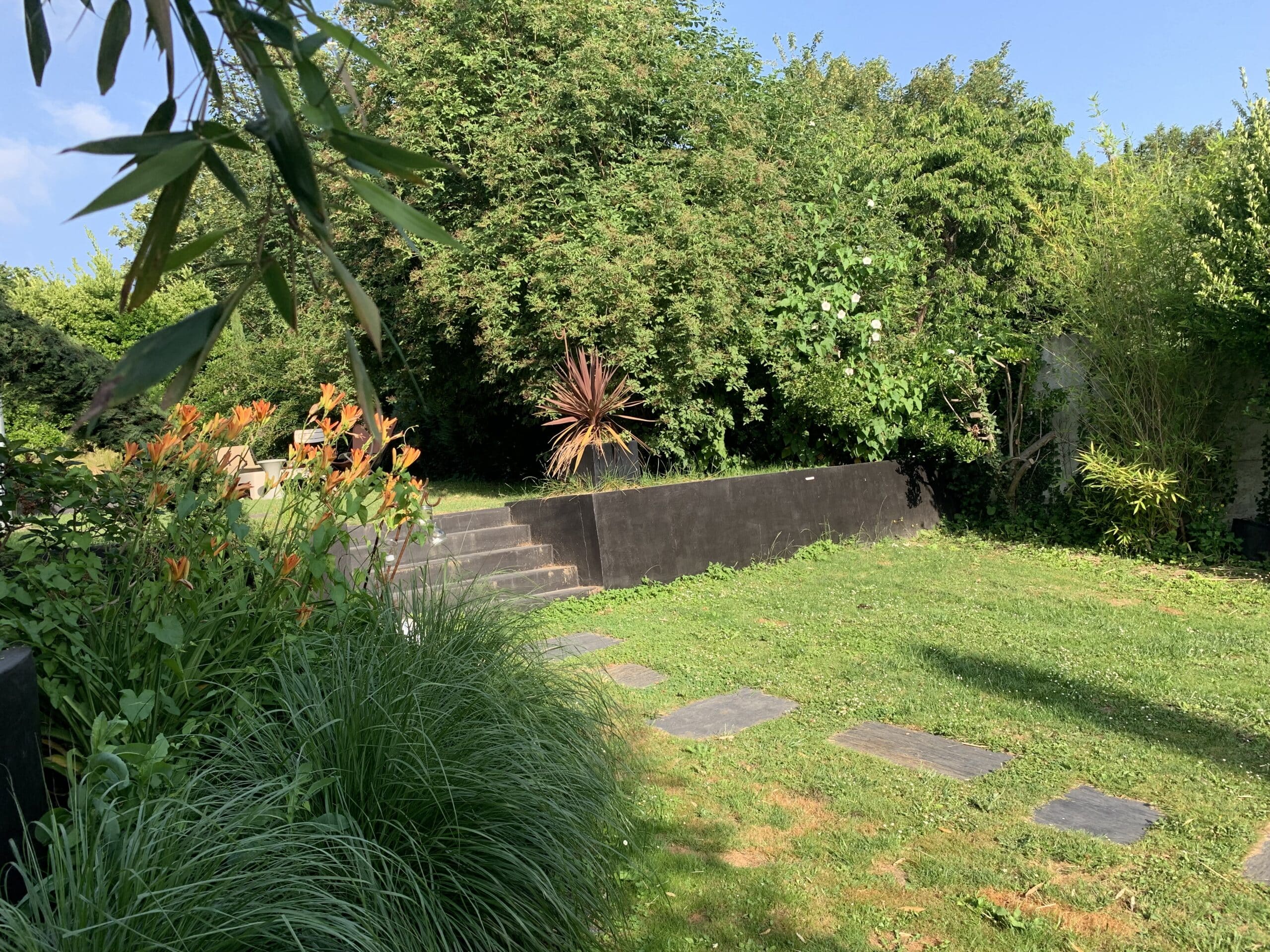 Jardin ensoleillé avec hémérocalles oranges, pelouse verdoyante, mur de soutènement noir, marches et plantes luxuriantes.