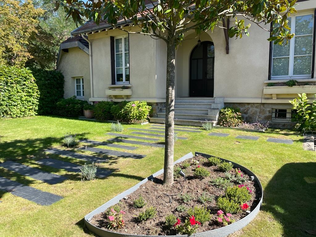 Maison beige avec porte arche, jardin verdoyant, pelouse, allée de pavés et parterre de fleurs colorées autour d'un arbre au soleil.