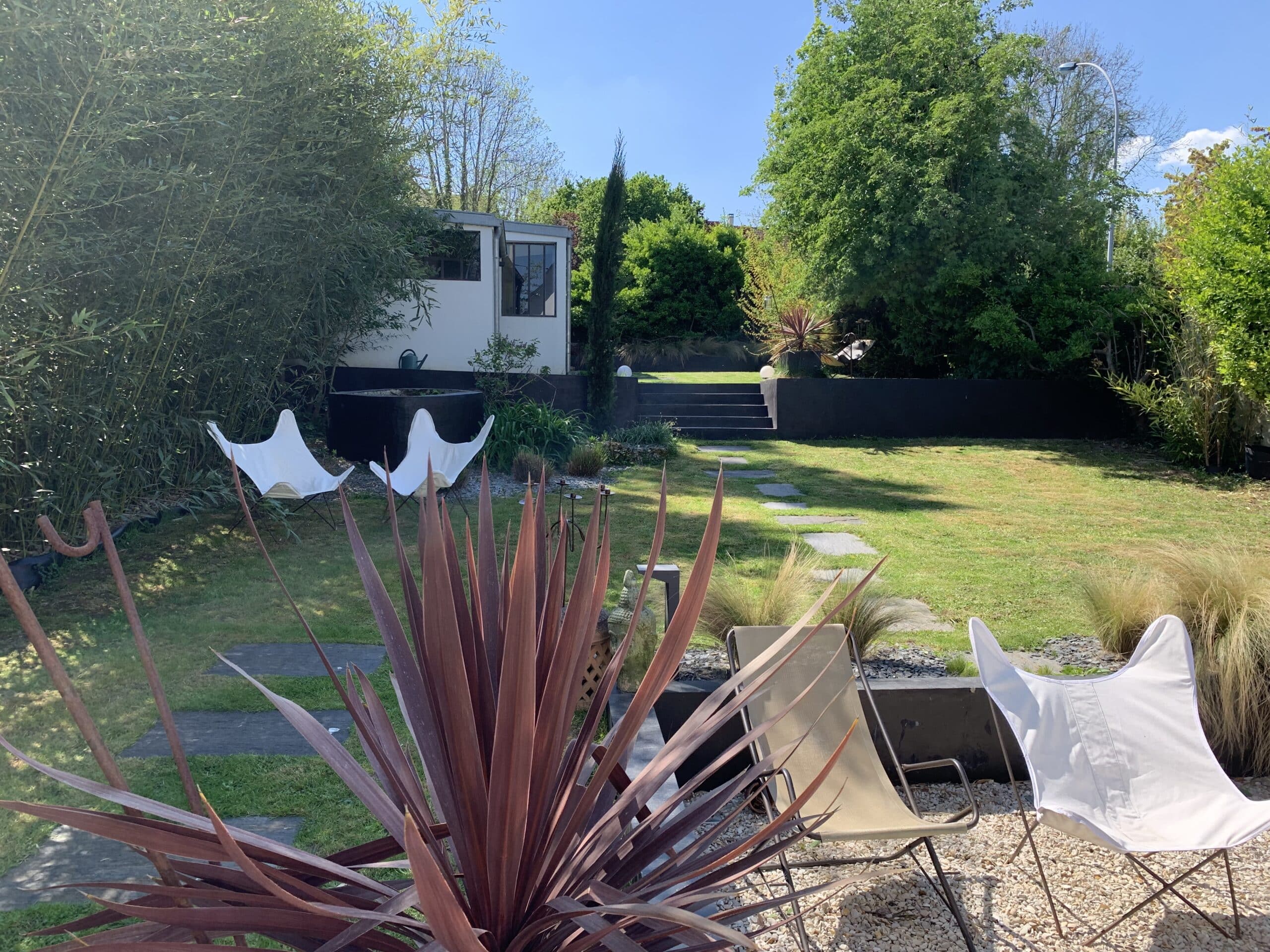 Jardin ensoleillé : haie de bambou, plantes, chaises papillon et transat. Allée de pierres, escalier vers une terrasse moderne.