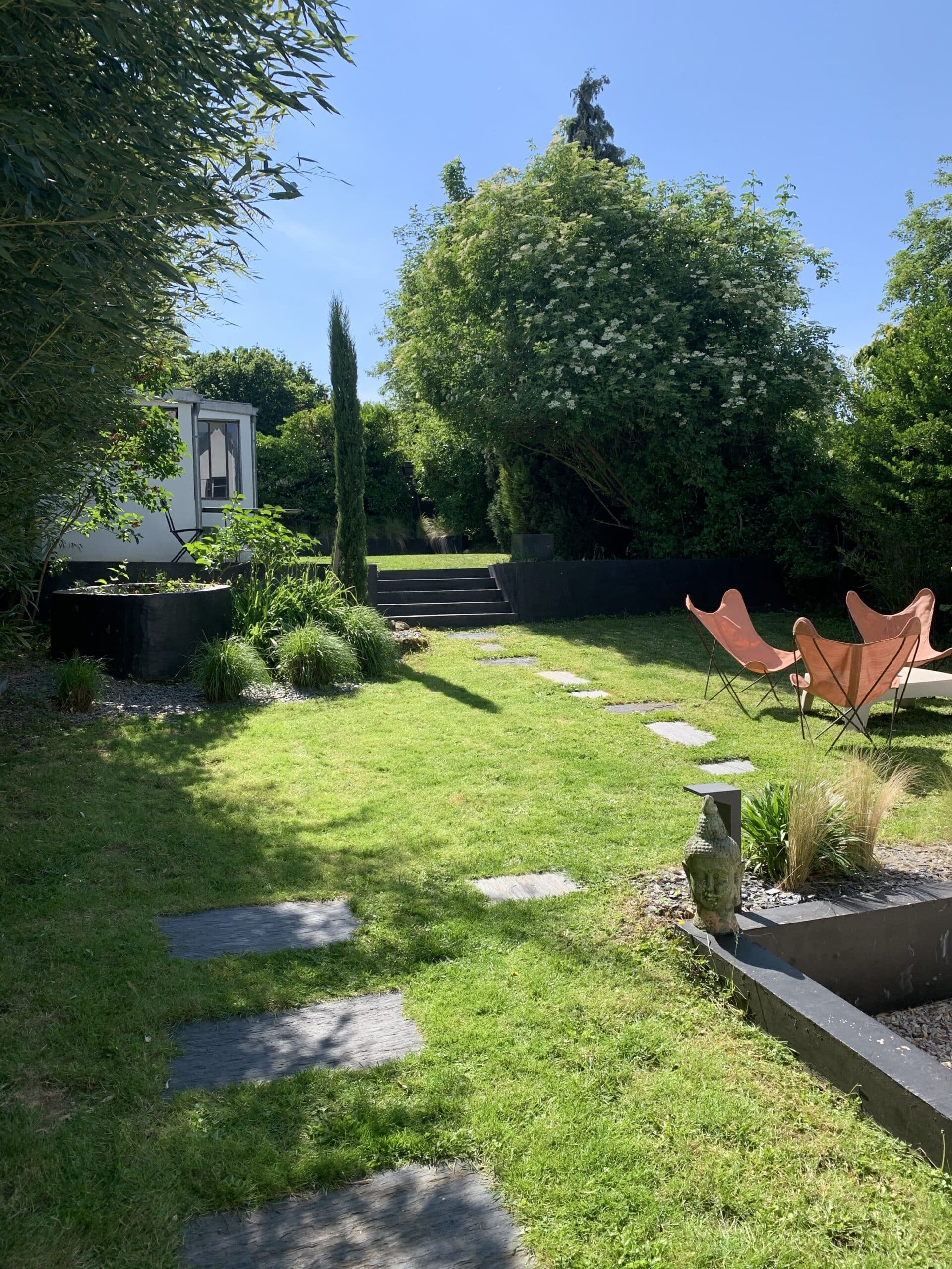 Jardin moderne et ensoleillé avec pelouse verte, chemin de dalles, cyprès, et bâtiment blanc. Deux chaises papillon orange et une tête de Bouddha ornent l'espace.