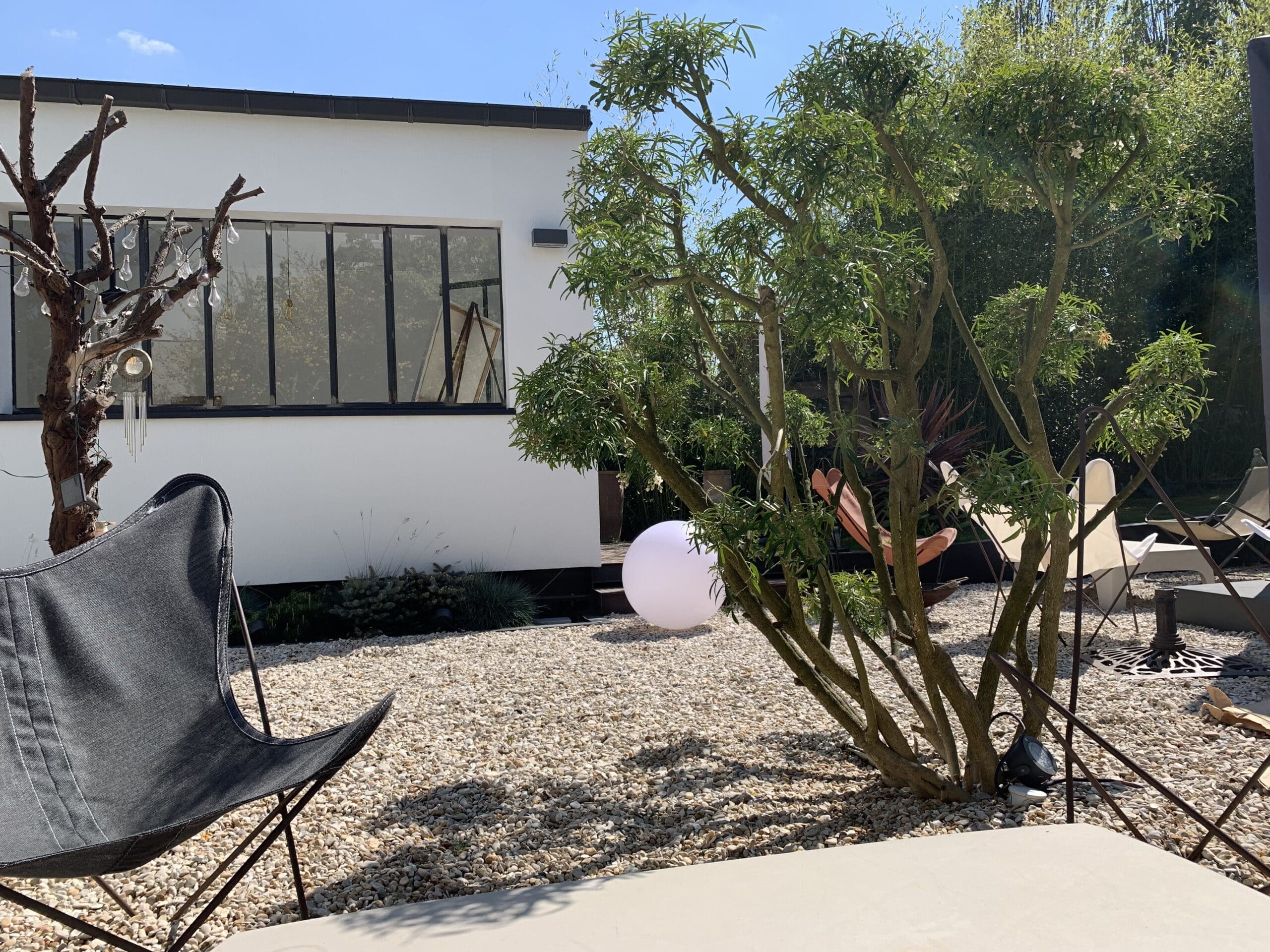 Jardin moderne avec chaise papillon noire, grand arbre fleuri, sol en gravier clair et maison blanche. Une lumière sphérique illumine l'espace.