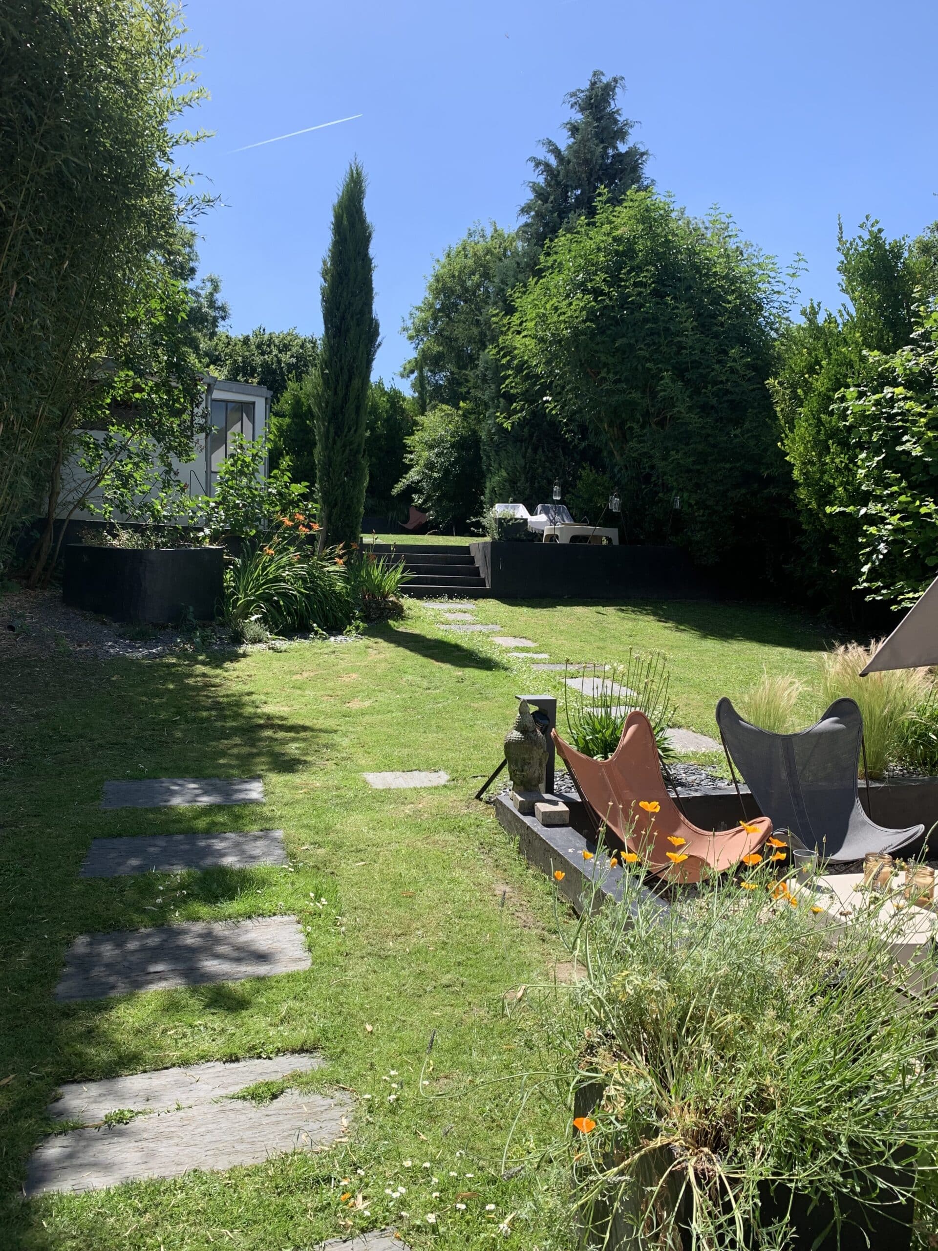 Jardin ensoleillé et luxuriant: chaises design, statue Bouddha, dalles sur pelouse verte, plantes fleuries et bâtiment. Ciel bleu, contrail d'avion.