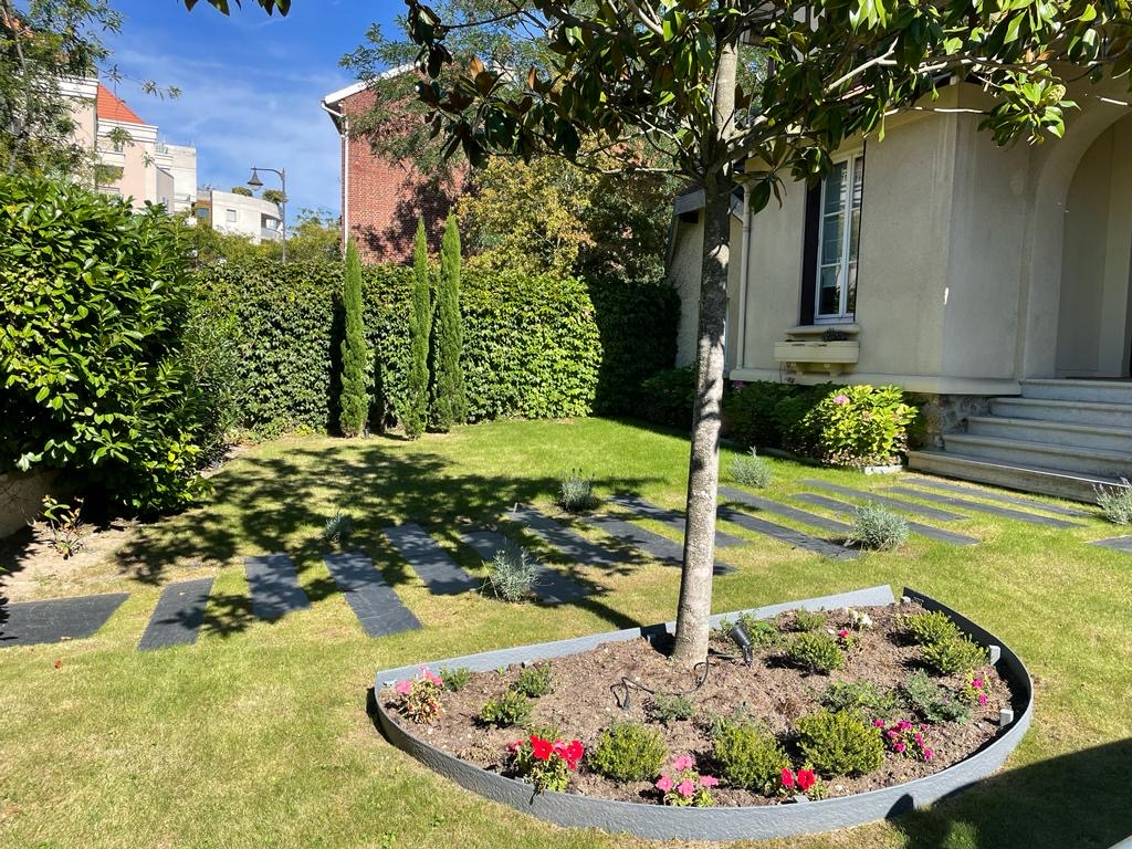 Jardin ensoleillé avec pelouse verte, massif fleuri autour d'un arbre, allée de dalles vers une maison claire. Haies luxuriantes et cyprès en arrière-plan.