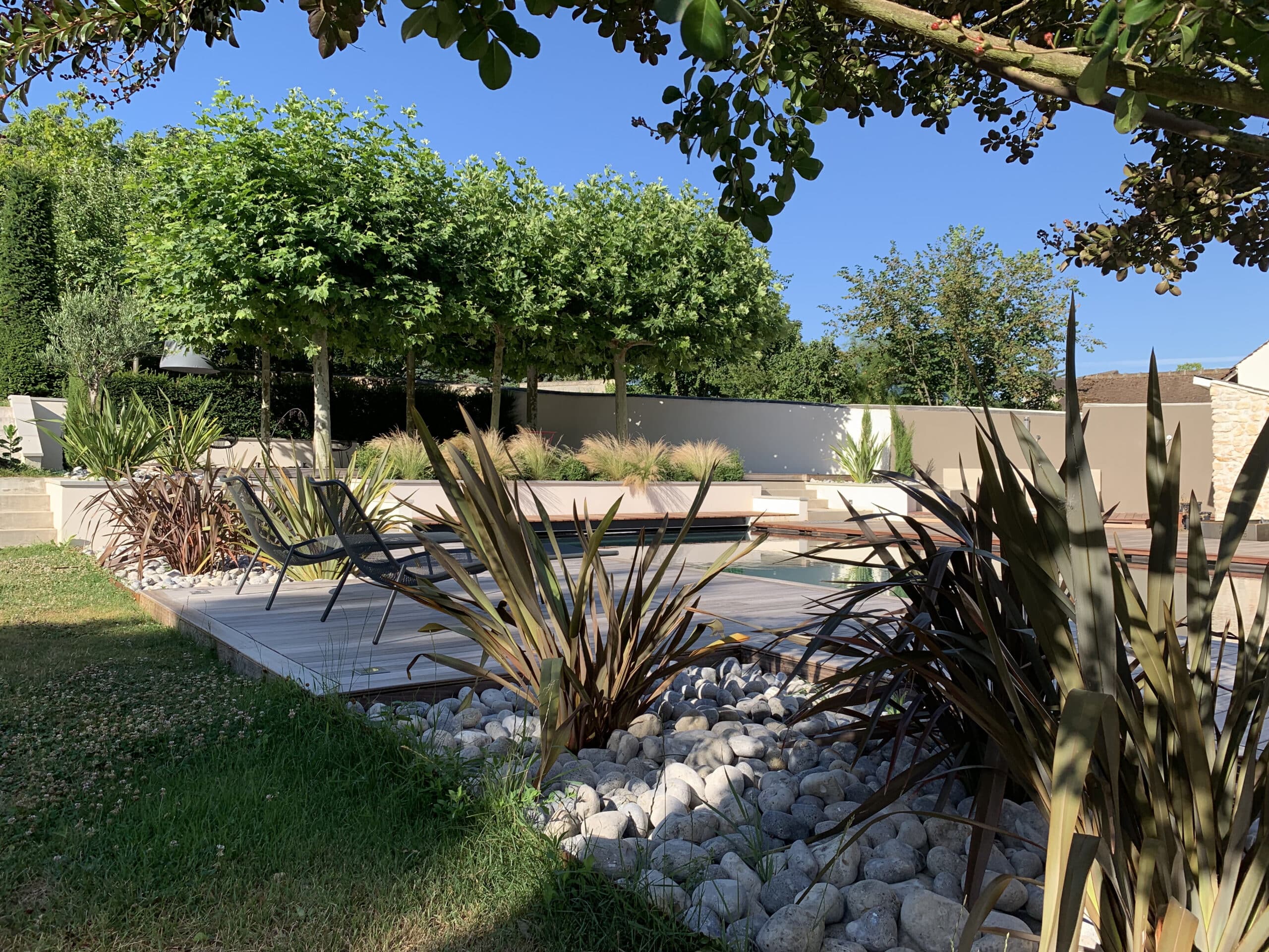 Jardin moderne avec piscine, terrasse en bois et mobilier design. Plantes exotiques, galets blancs et arbres luxuriants sous un ciel bleu clair.