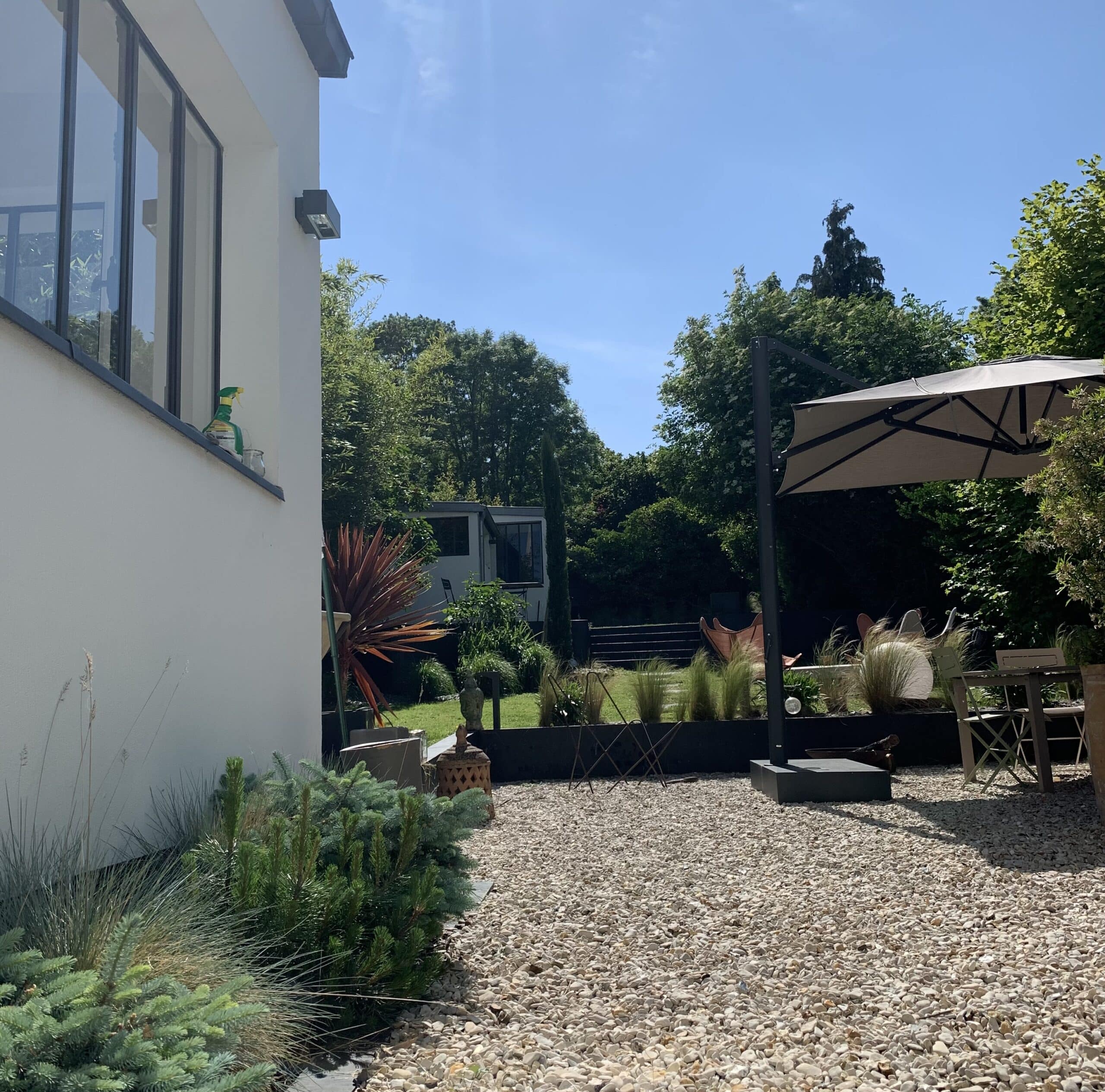 Maison moderne lumineuse, grandes fenêtres. Jardin de gravier luxuriant, parasol beige, mobilier extérieur sous ciel bleu ensoleillé.
