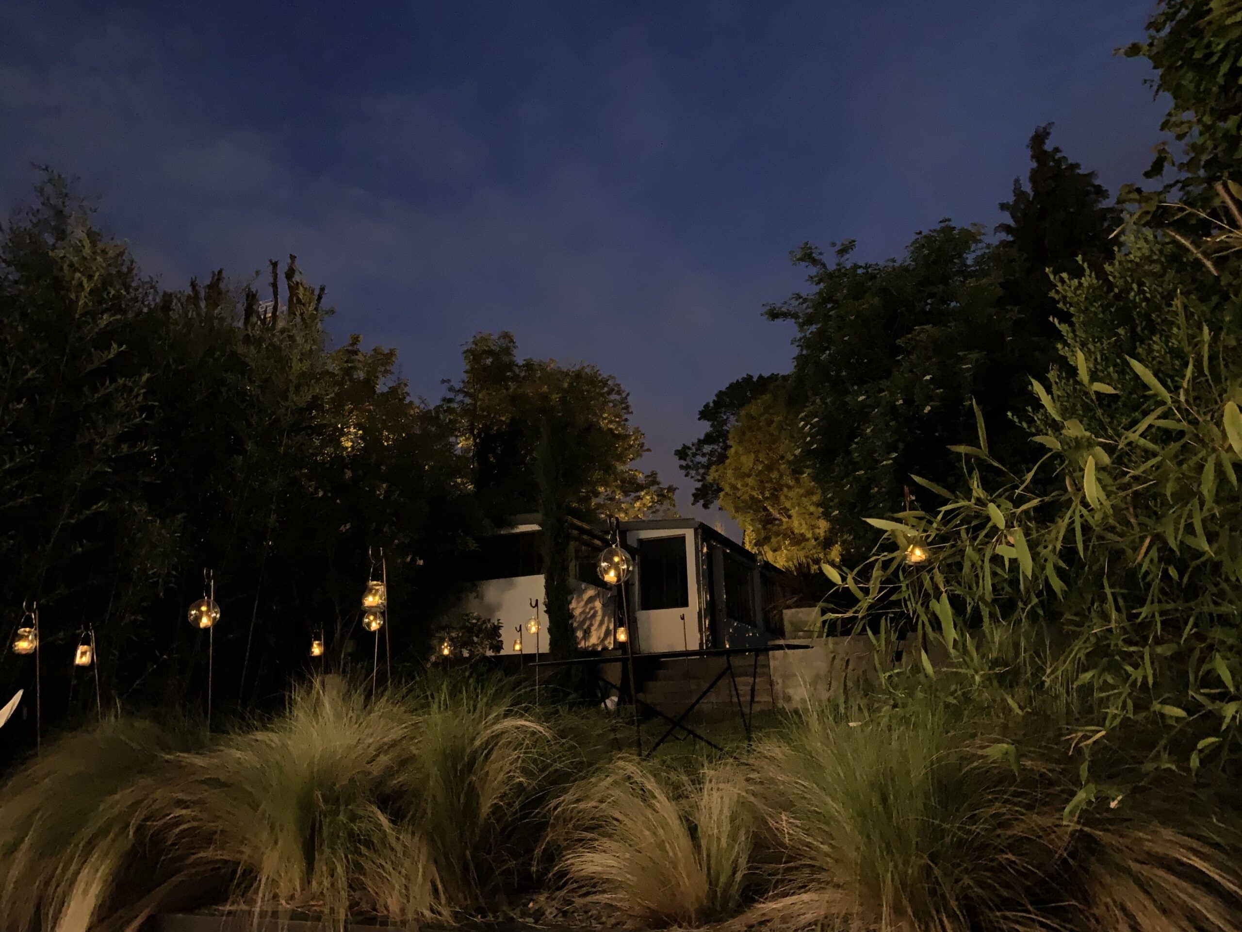 Jardin de nuit: lanternes lumineuses parmi des graminées, arbres et bâtiment moderne sous un ciel sombre.