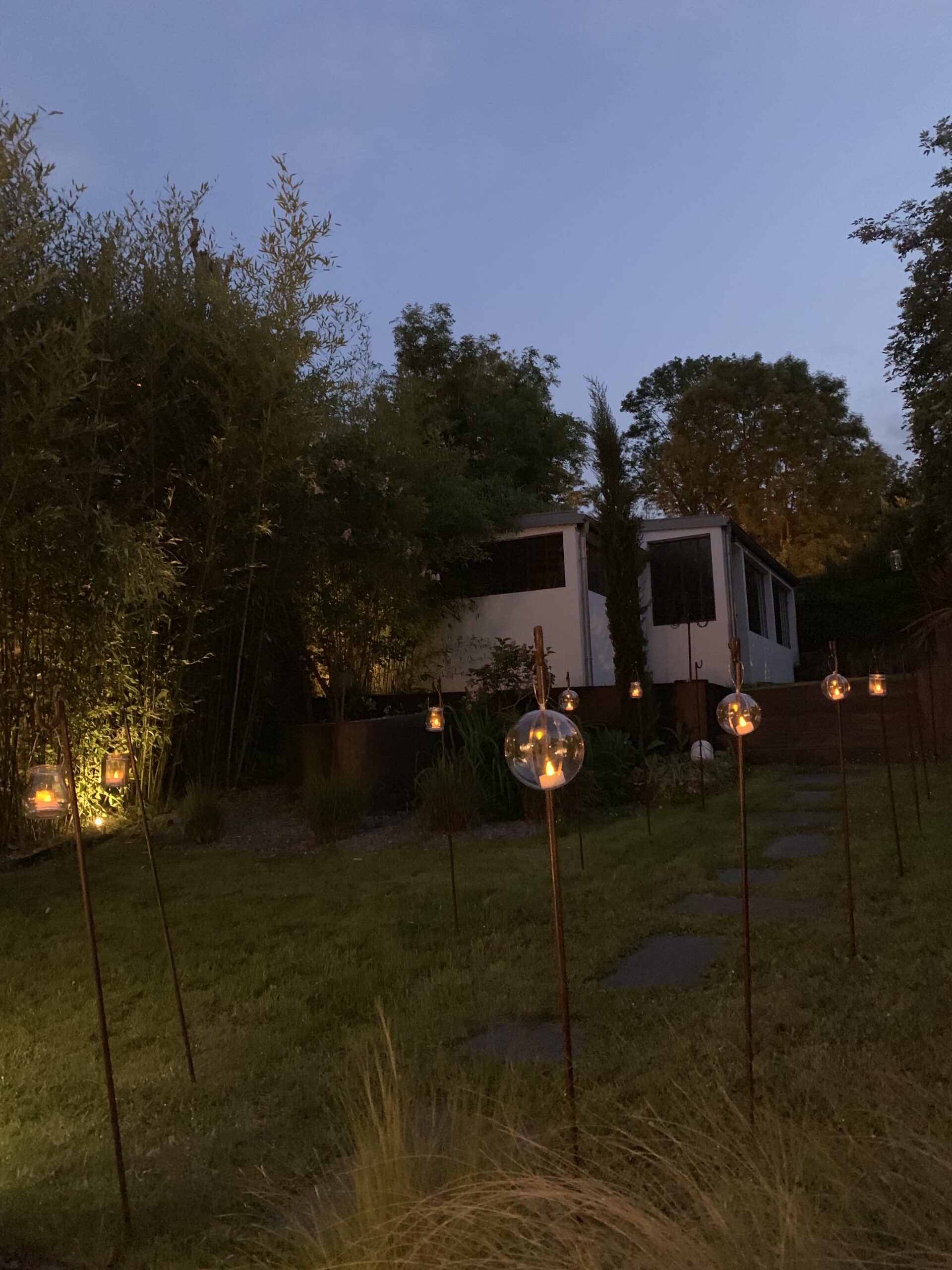 Jardin illuminé la nuit avec des lanternes suspendues et des orbes lumineuses sur des piquets, bordé de bambous luxuriants et d'une maison lointaine.